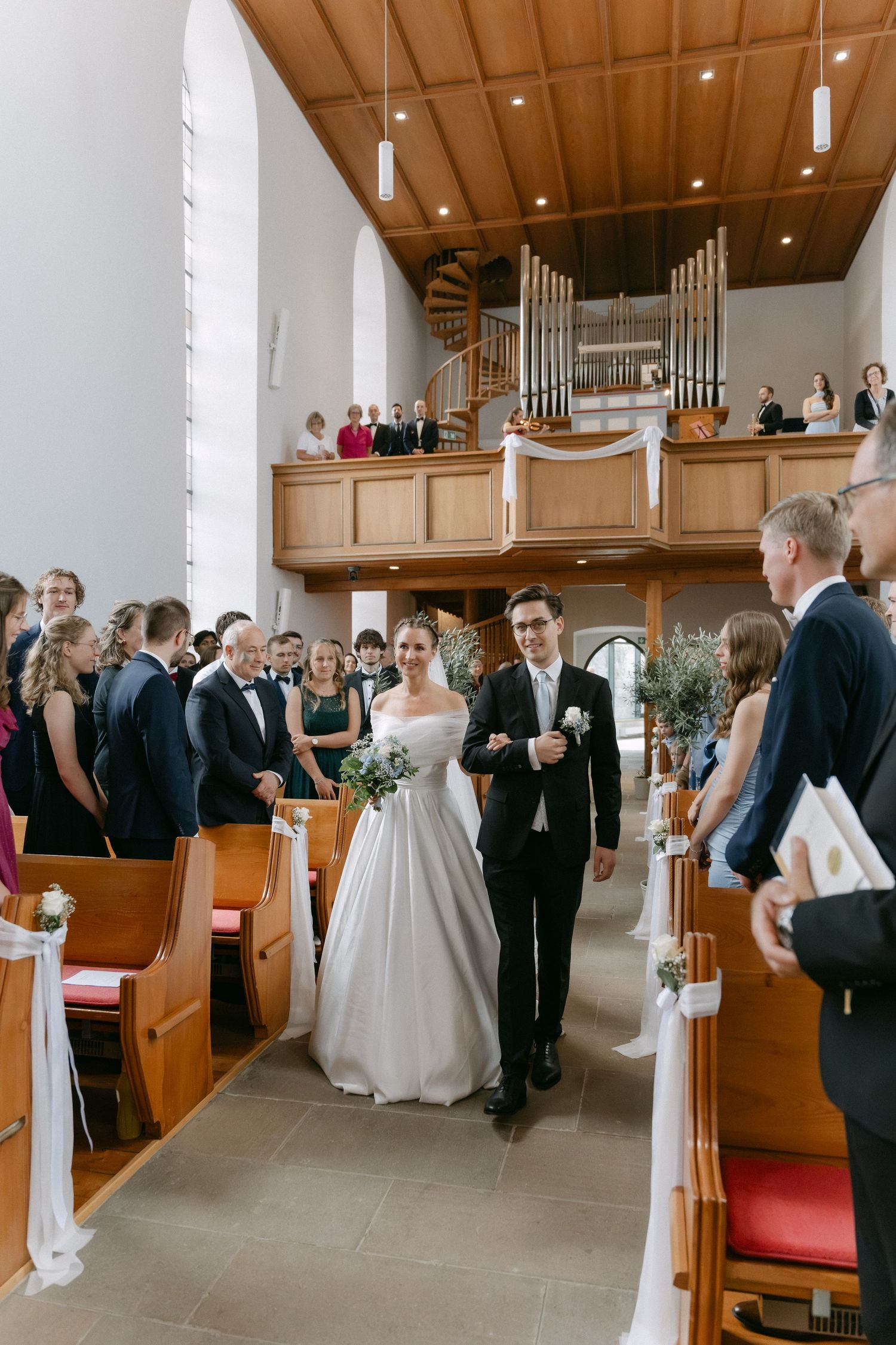 Bride in a white gown holding a bouquet walks down the church aisle with the groom as smiling wedding guests look on.
