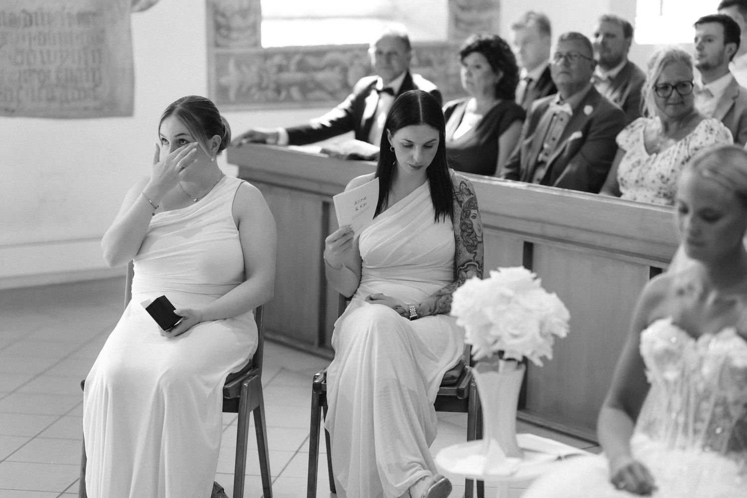Black and white photo of a bridesmaid wiping a tear during a wedding ceremony while guests sit in the background.