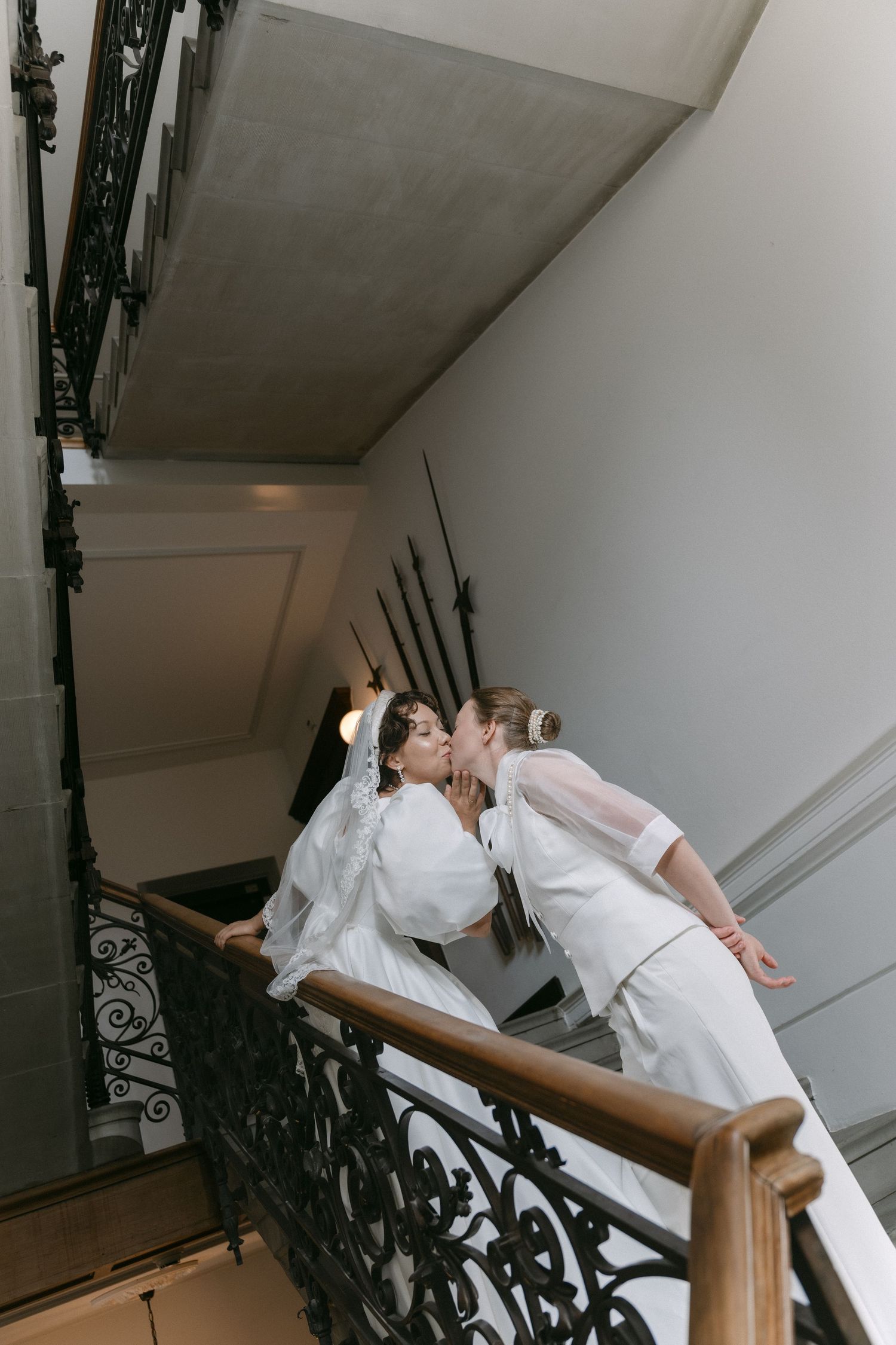 Two brides in white gowns share a kiss on an elegant staircase with ornate iron railings.