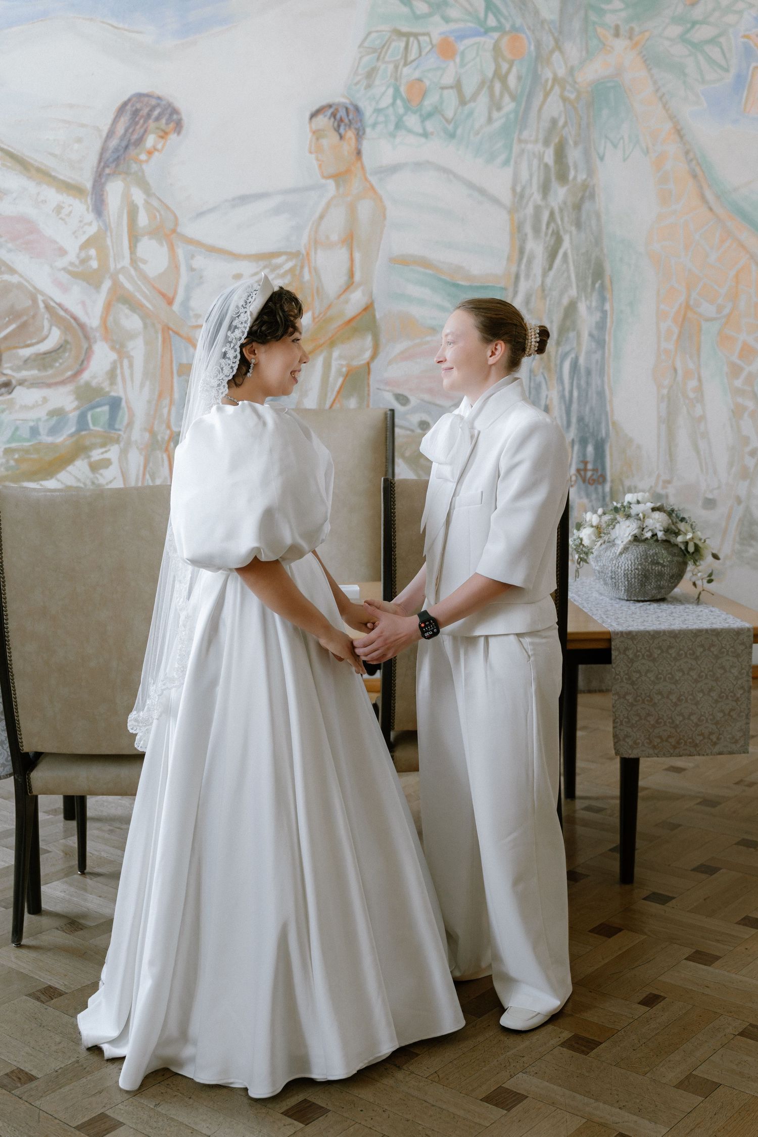 Two brides in white attire hold hands smiling at their wedding ceremony before a painted mural backdrop.