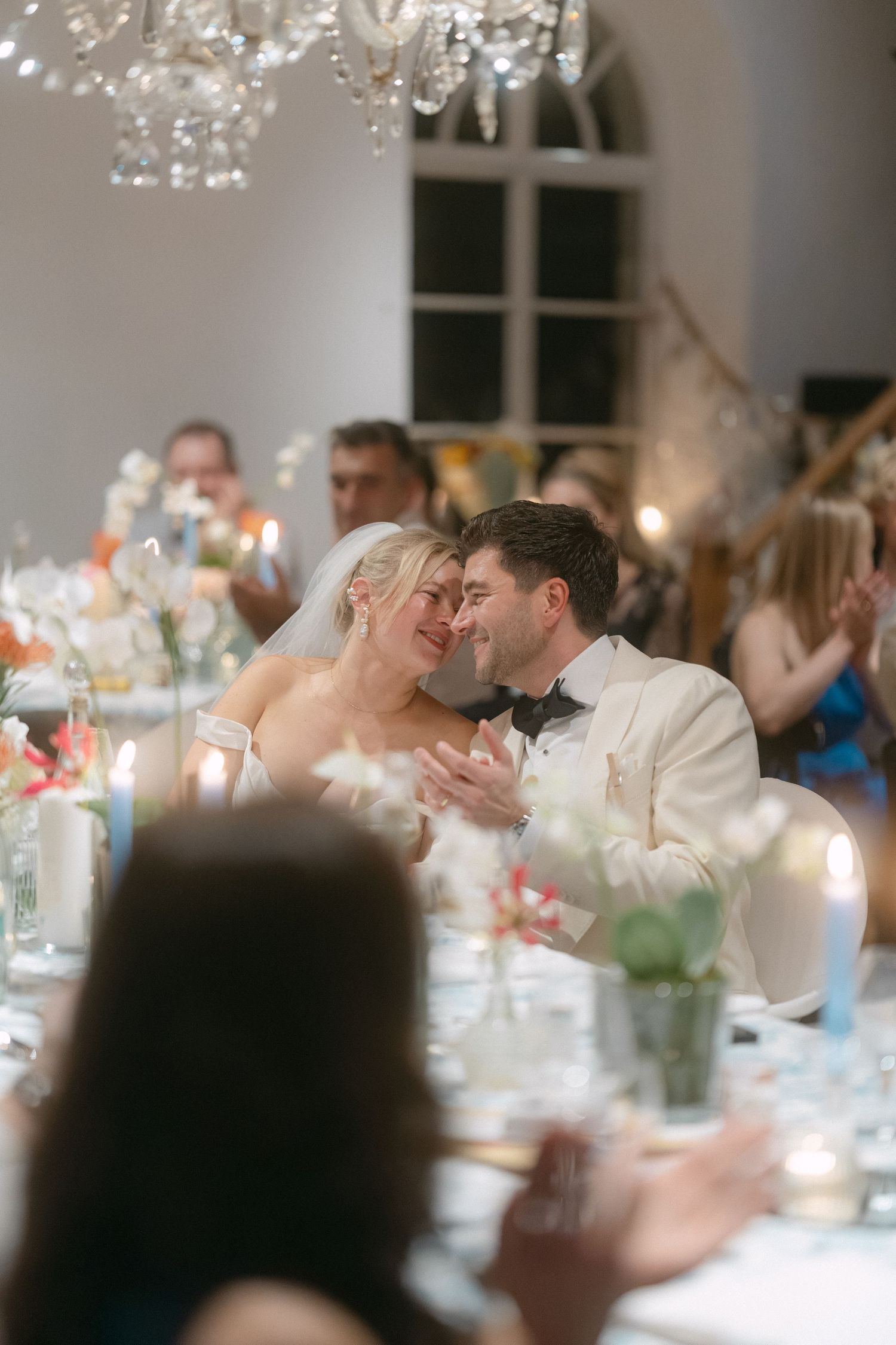 A romantic wedding reception moment captured at an elegant candlelit dinner table under a crystal chandelier.