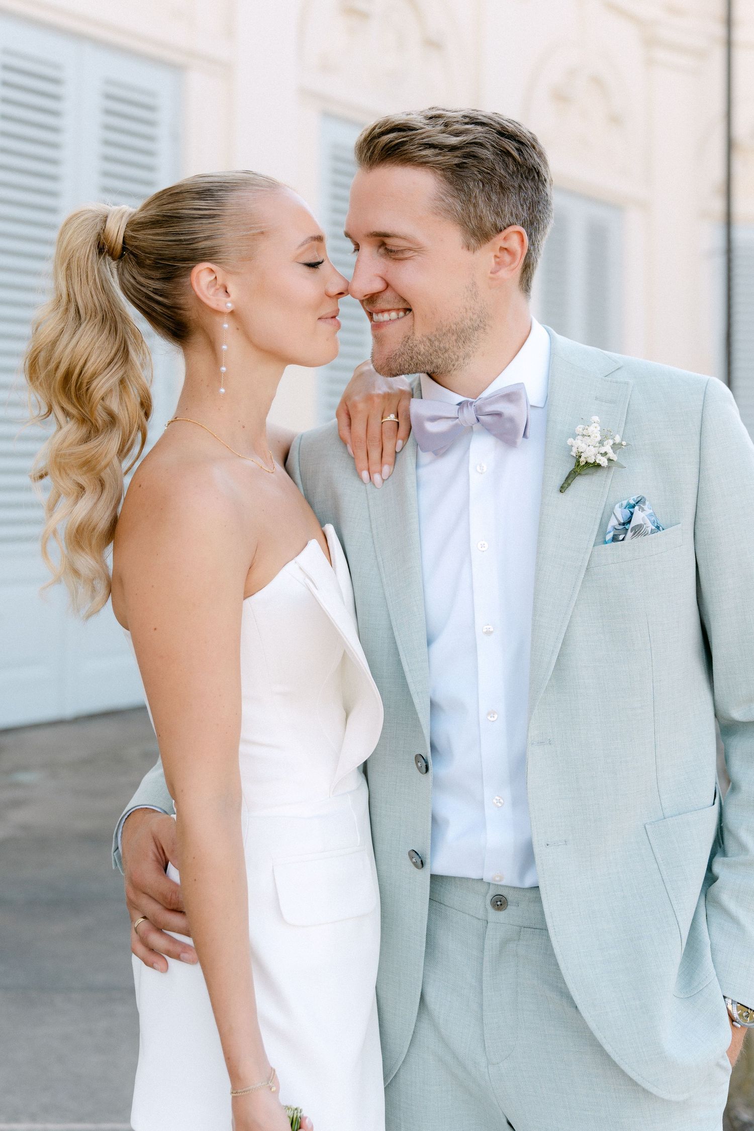 A romantic wedding couple in elegant attire share an intimate moment against a light blue wall with shutters. Schloss Solitude