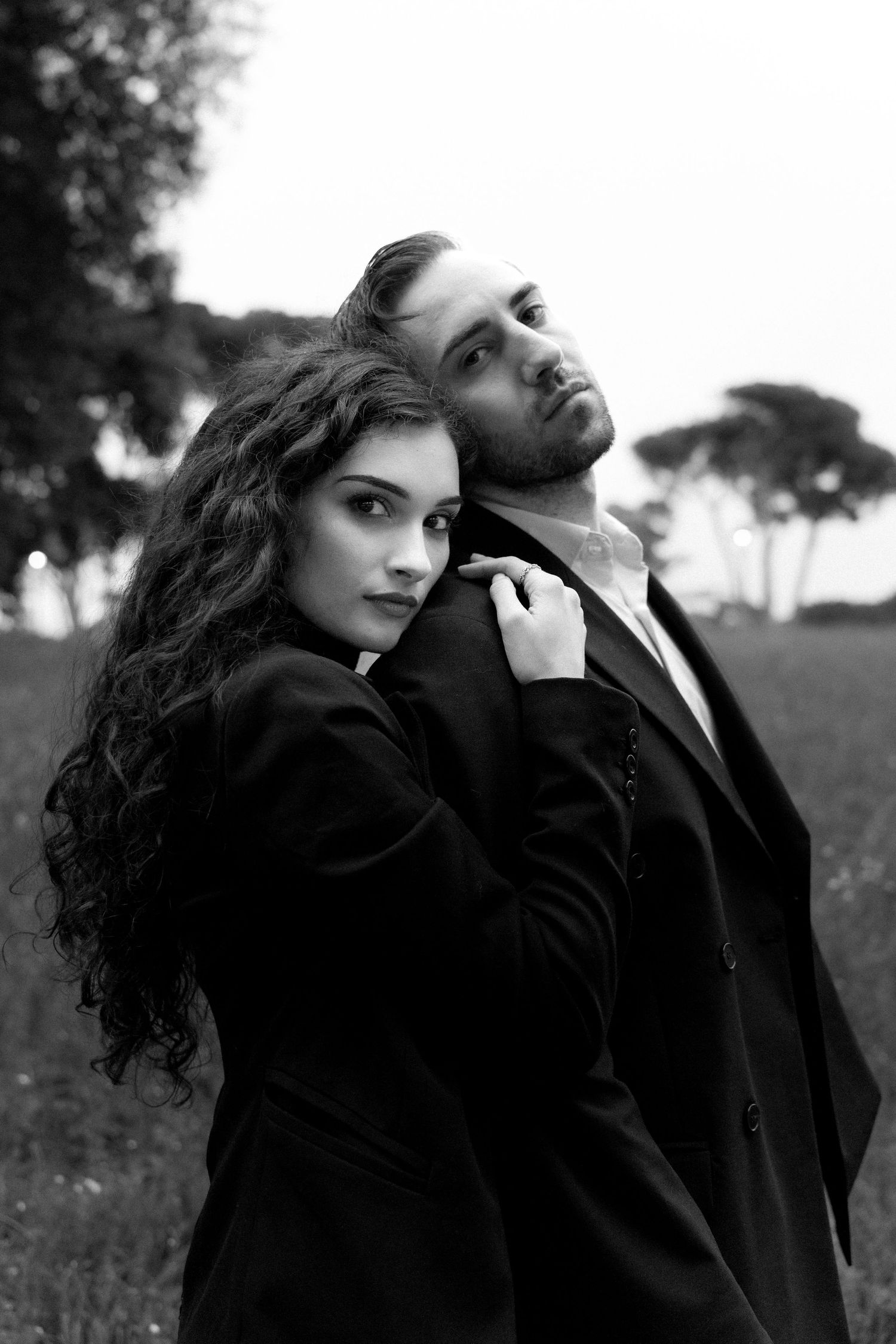 A dramatic black and white portrait of a couple in dark formal attire embracing while standing in a grassy field.