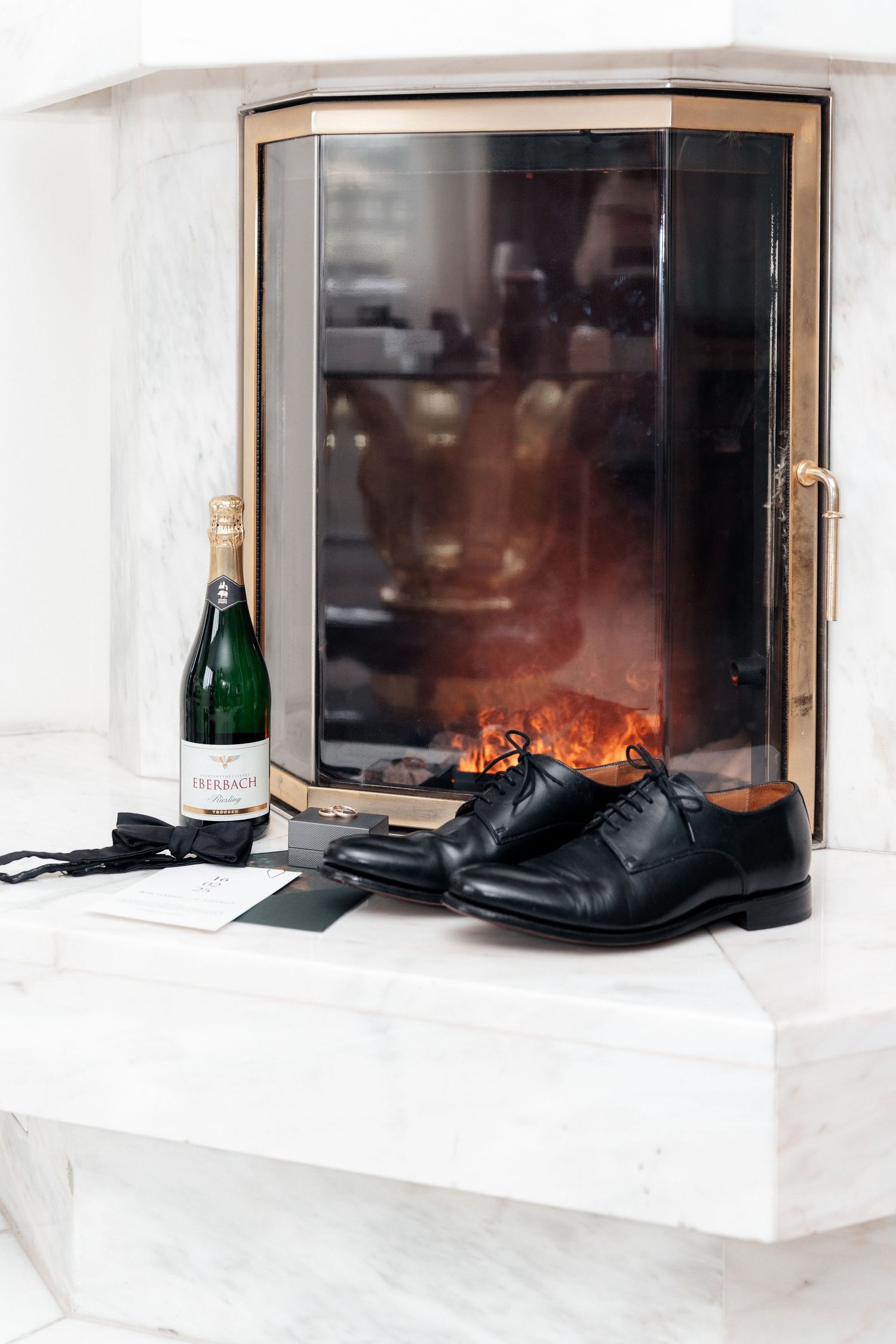 A pair of black dress shoes and champagne bottle sit on a white ledge next to a window with glowing firelight.