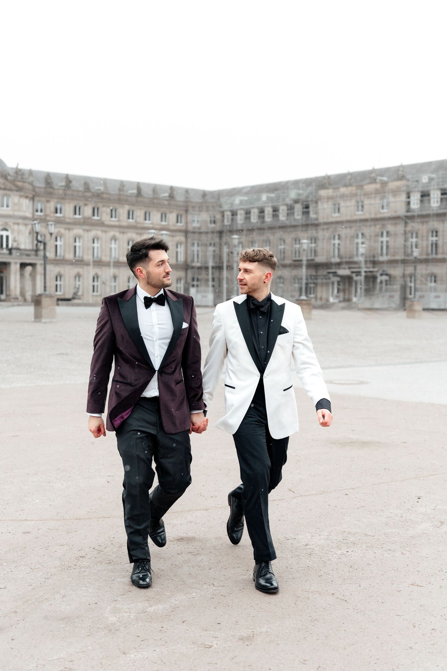 Two people in formal attire walk together outside a grand historic European palace courtyard.