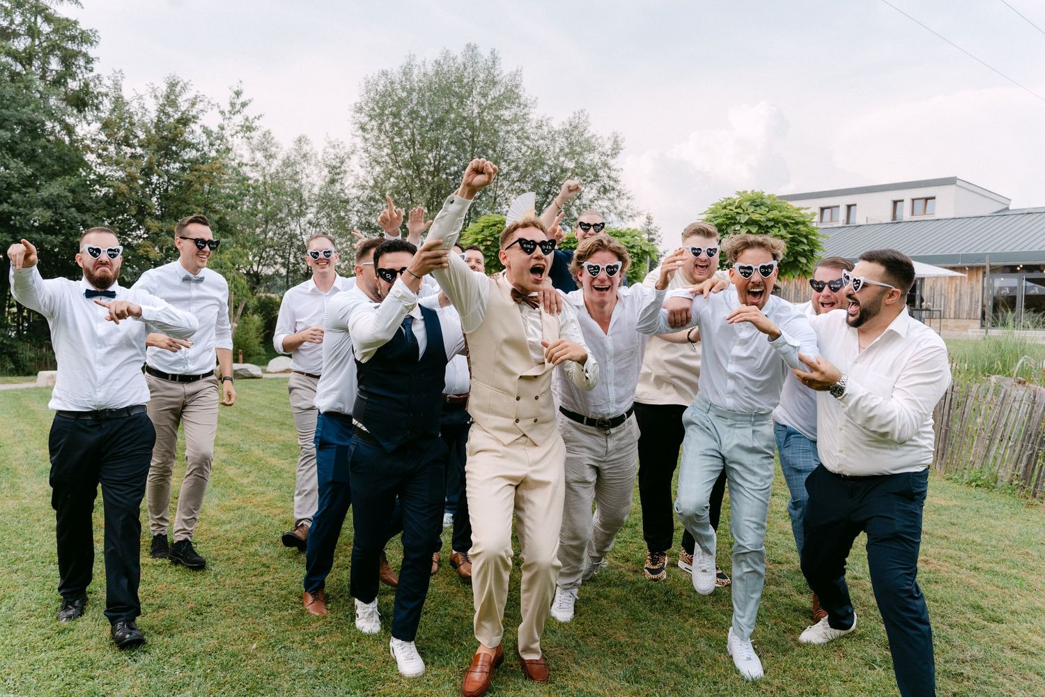 Groomsmen in light colored suits jumping and posing playfully on a grassy lawn at a wedding celebration.