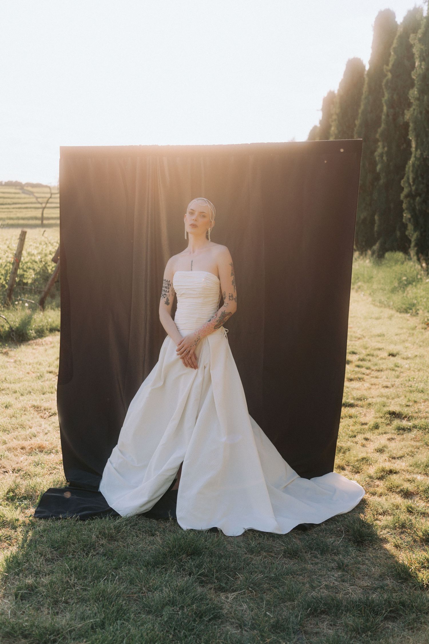 A figure in a flowing white wedding gown poses against a dark backdrop with sunlit fields in the background.