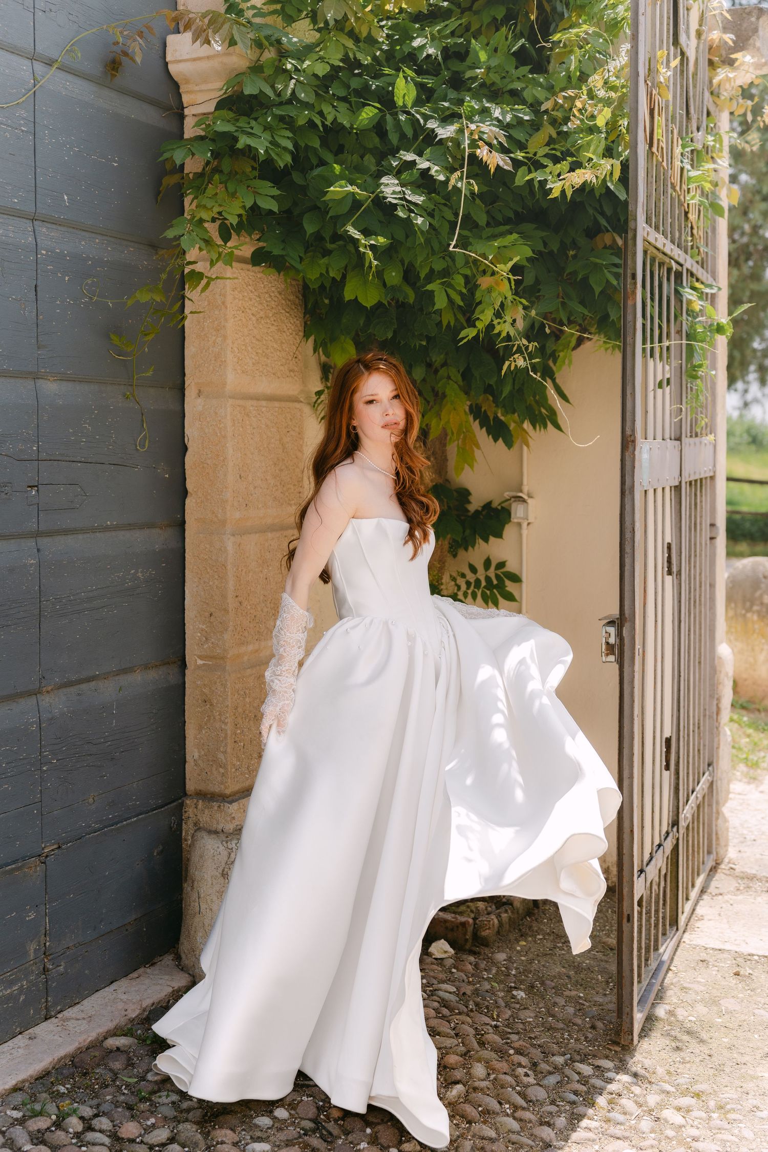 A bride in a flowing white wedding dress twirls gracefully in front of a stone wall with green foliage overhead.