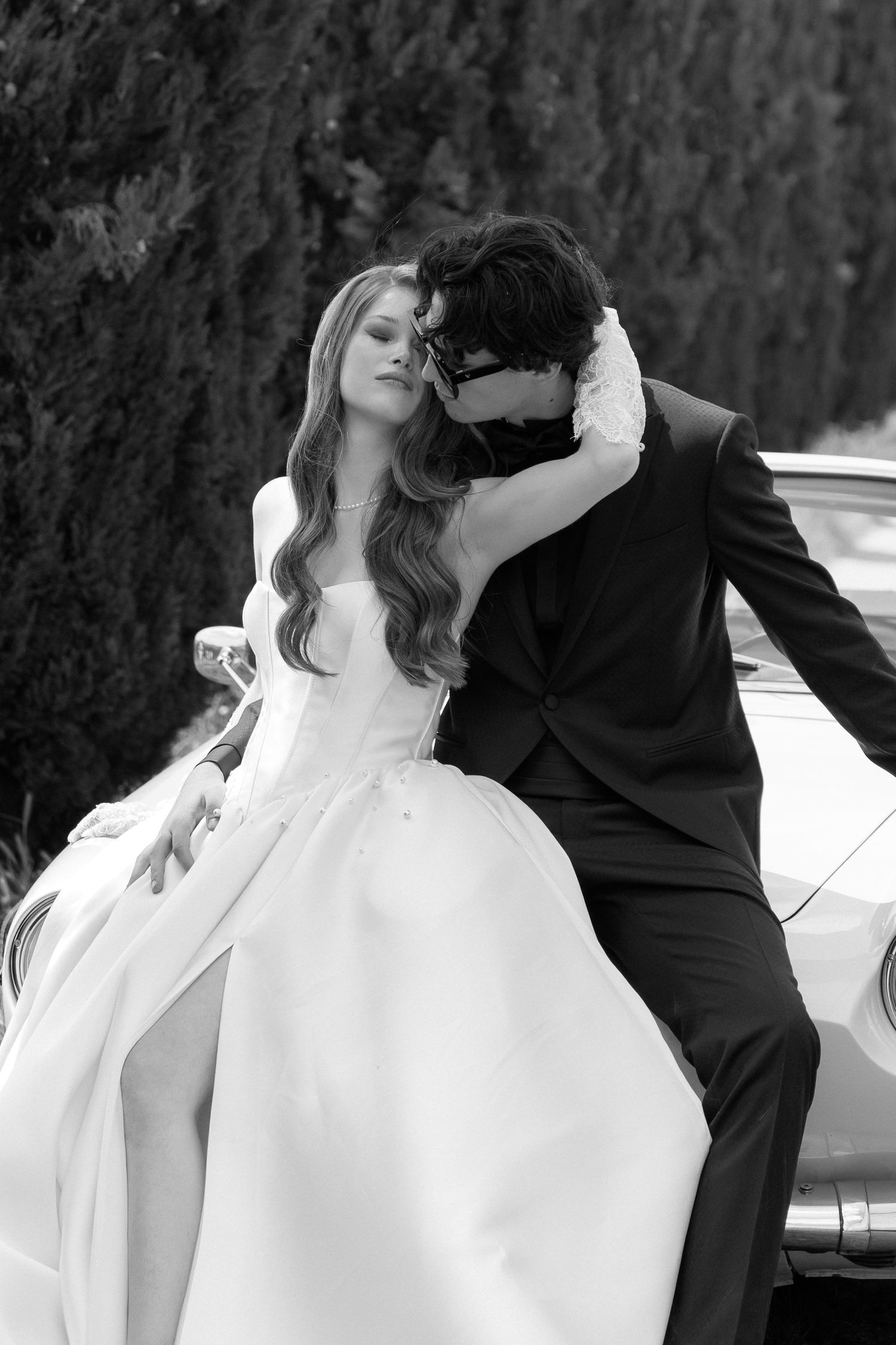 A romantic black and white wedding photo of a couple embracing near a vintage car, with the bride in a flowing white gown.