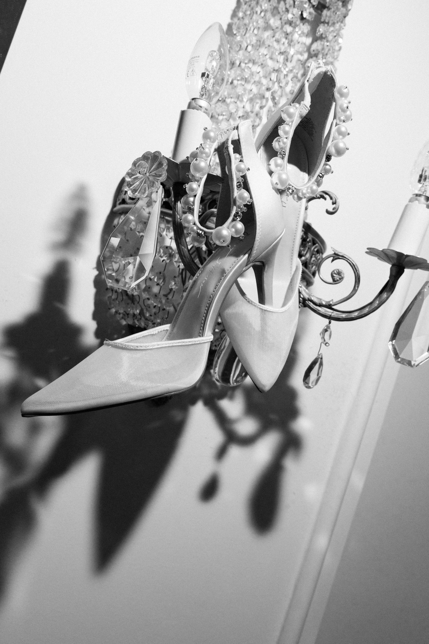 Elegant white high heel shoes displayed on a crystal chandelier casting dramatic shadows in black and white photography.