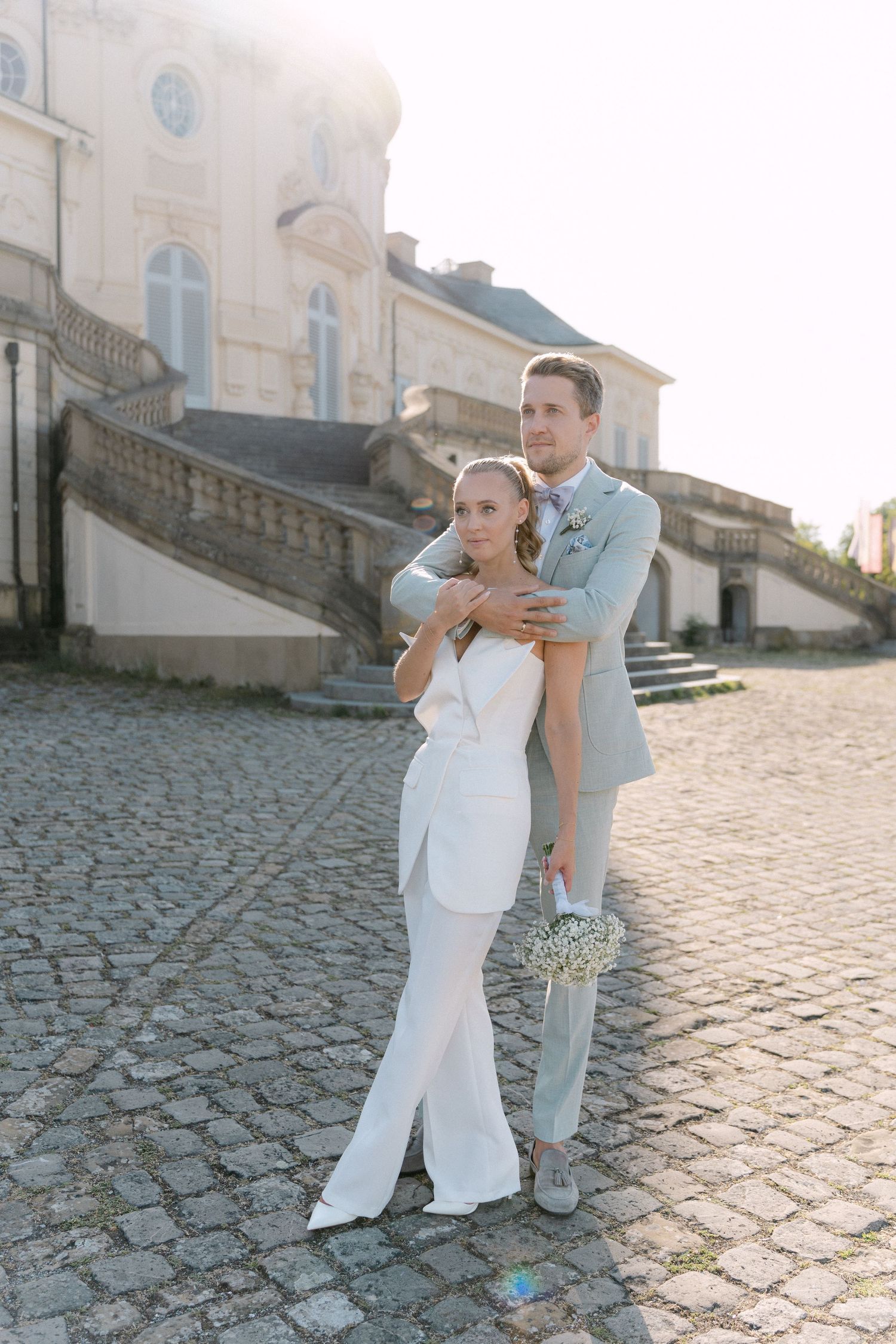 A romantic wedding couple embraces on cobblestone steps outside an elegant historic building on a sunny day. Schloss Solitude