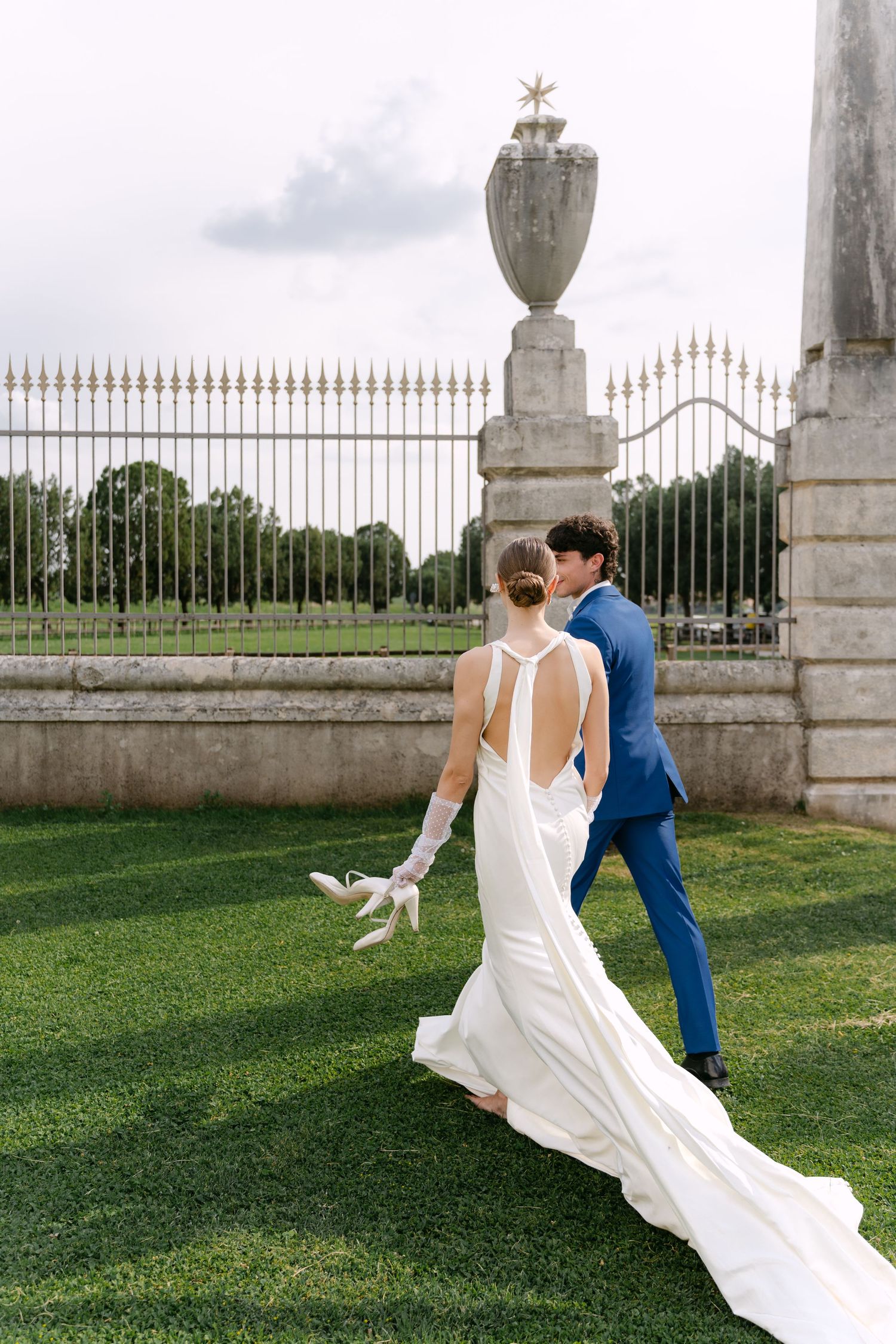 A couple in wedding attire walk on manicured lawn near an ornate stone fence with decorative urn at an elegant venue. Villa Mosconi Bertani