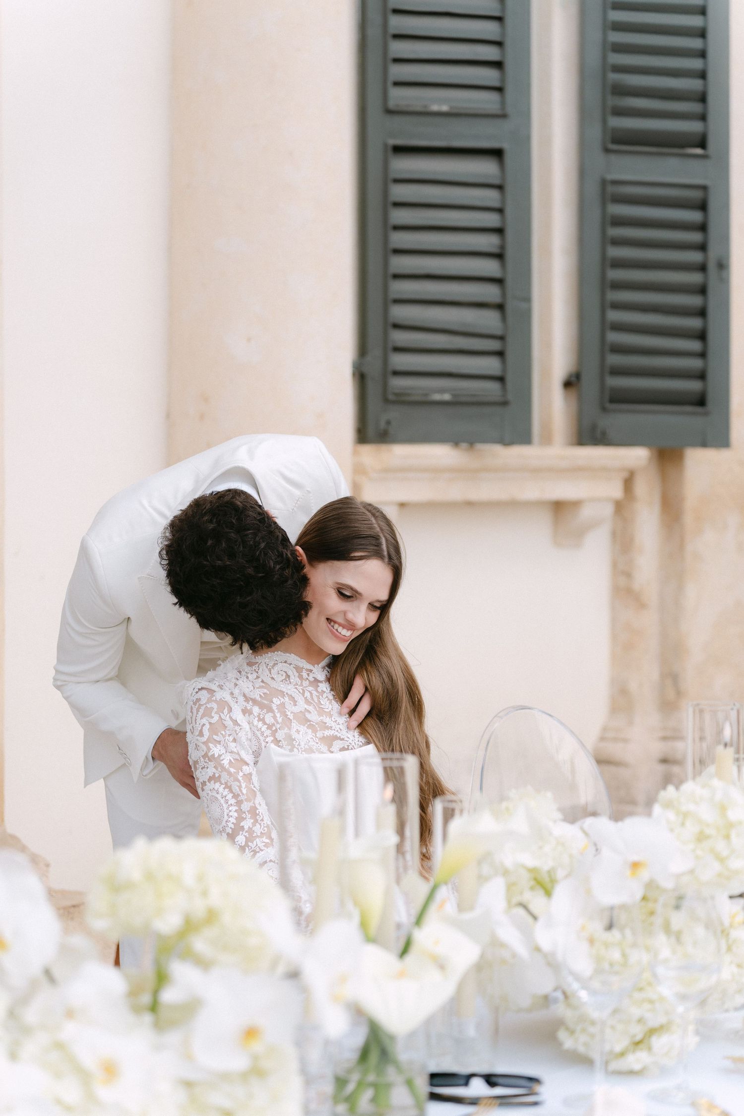 A bride in a white lace dress leans over an elegantly set table with white floral centerpieces. Villa Mosconi Bertani
