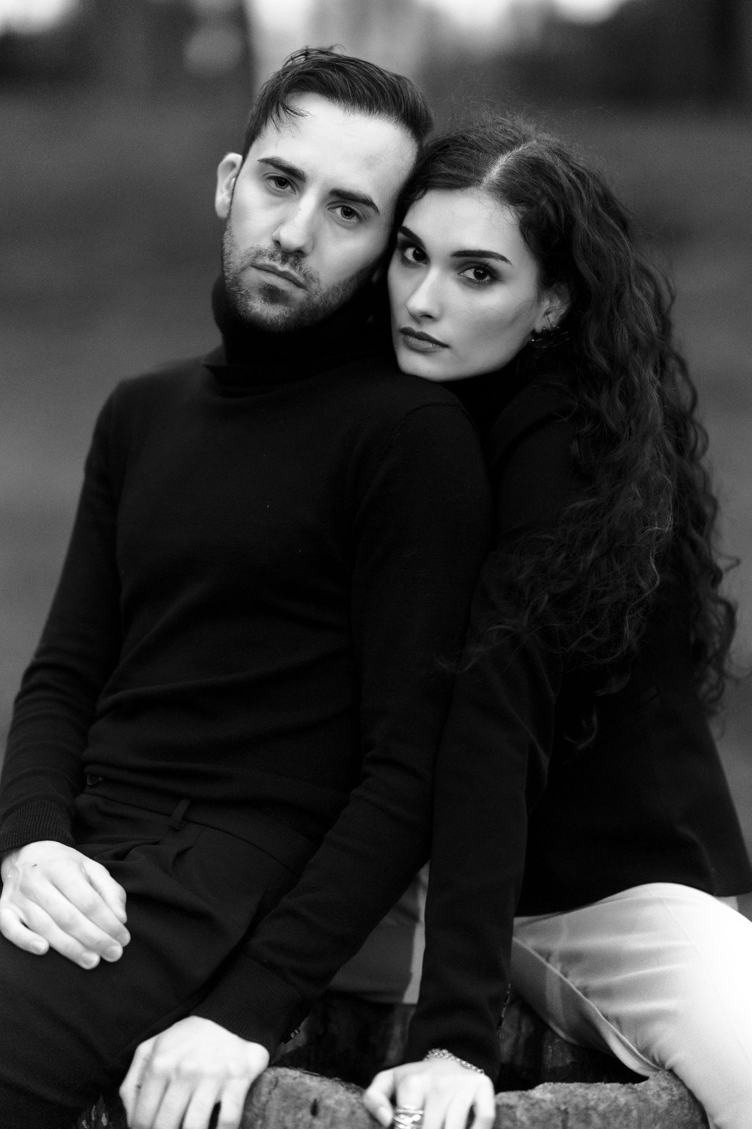 Black and white portrait of a couple in dark sweaters posing intimately against blurred background.