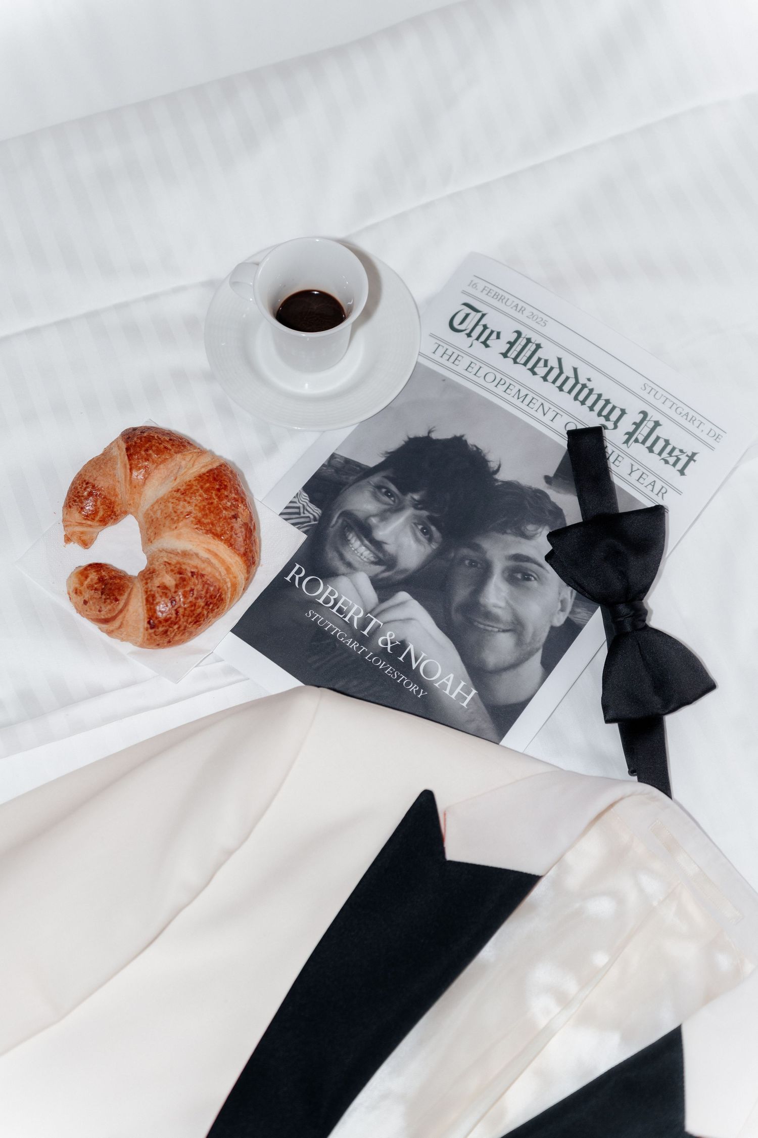 Flatlay with coffee cup, croissant, newspaper with black ribbons, and white fabric on light surface.