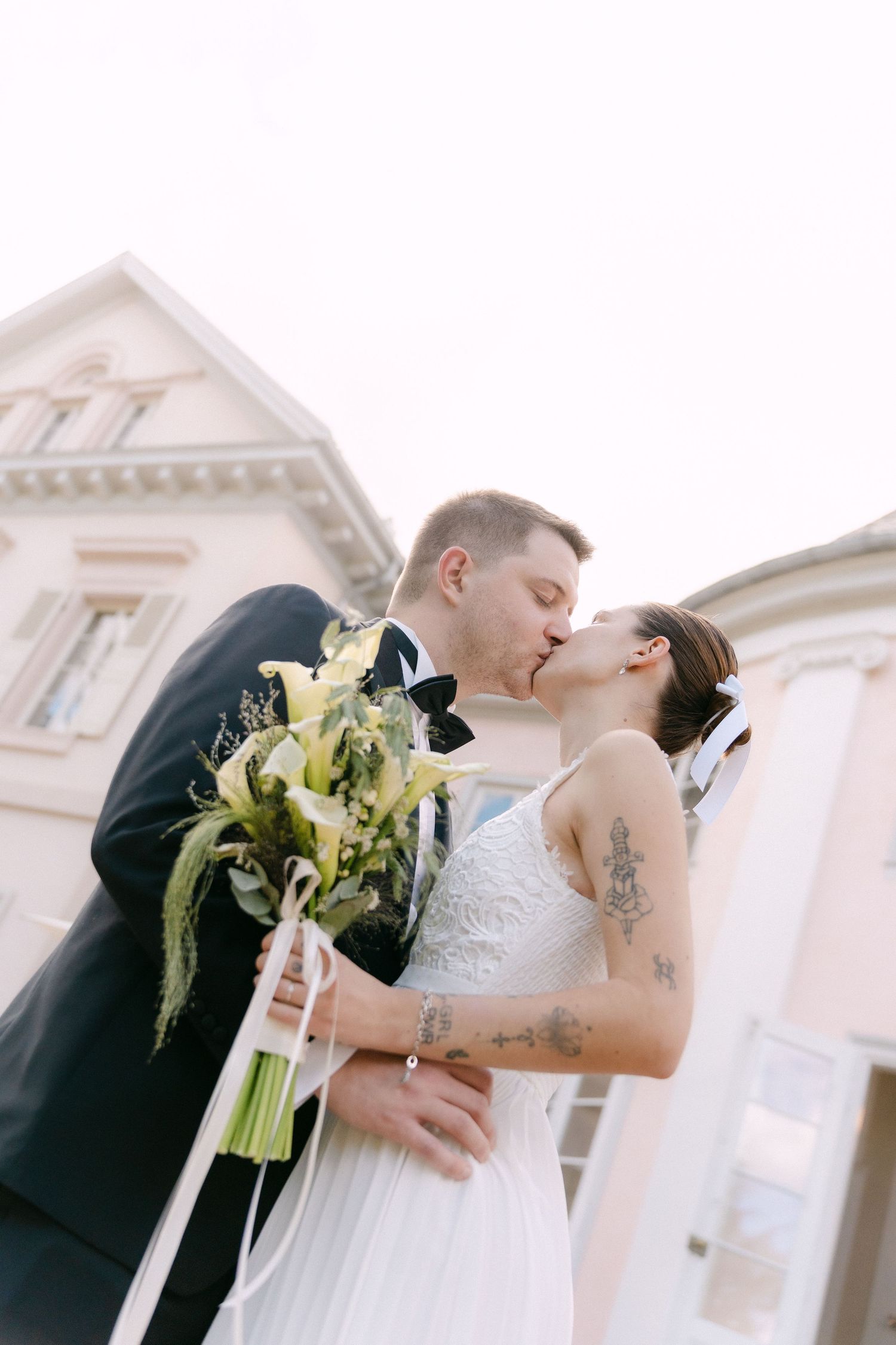Bride and groom sharing a romantic kiss outdoors in wedding attire, bride holding white lily bouquet with ribbon.
