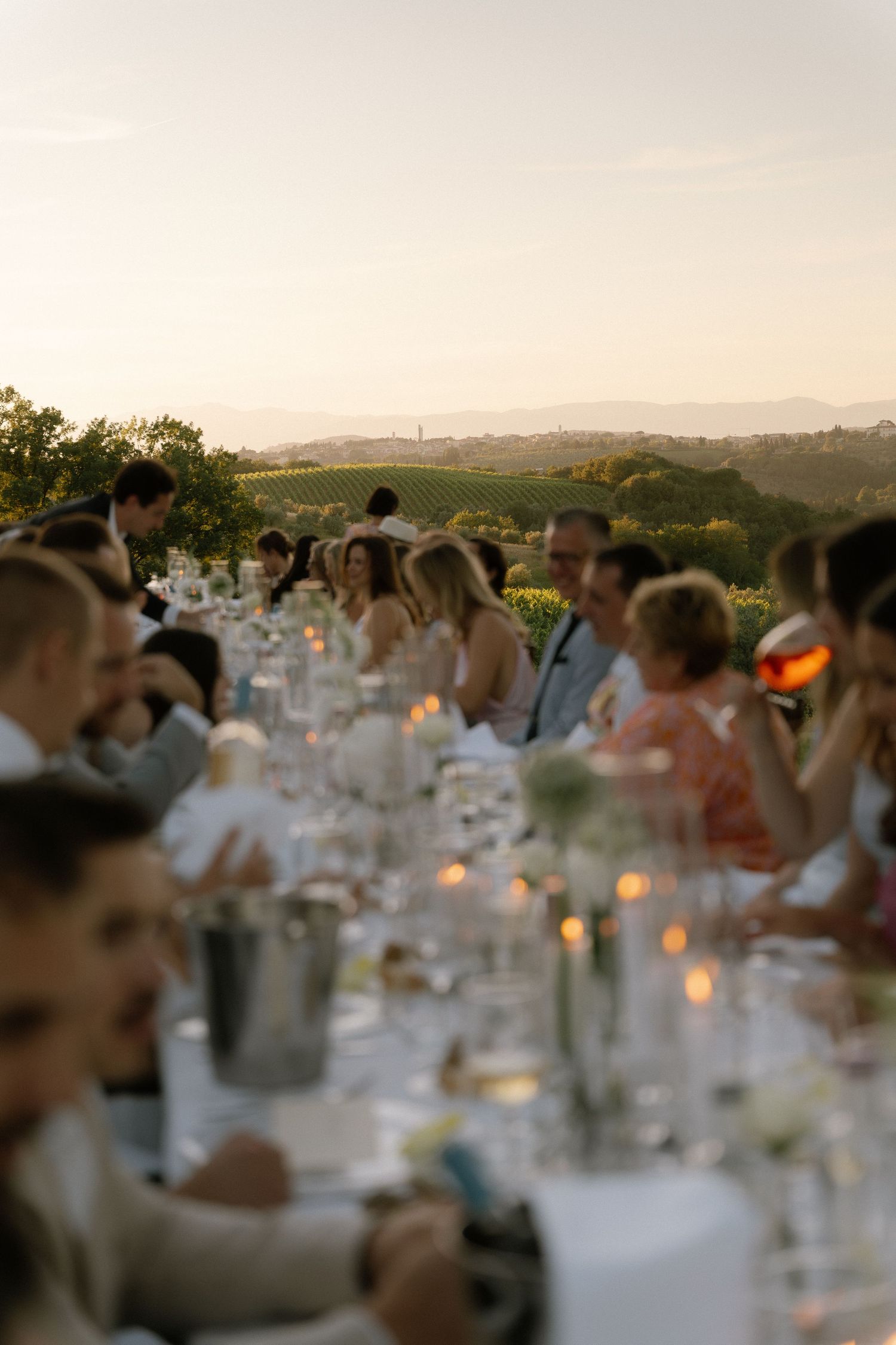 Elegant outdoor wedding reception dinner at sunset with long candlelit table, guests in formal attire, vineyard backdrop.