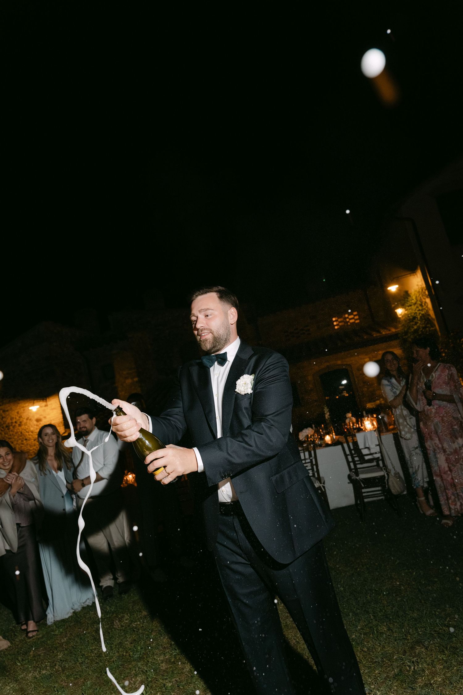 Groom in black tuxedo popping champagne bottle at outdoor wedding reception at night.