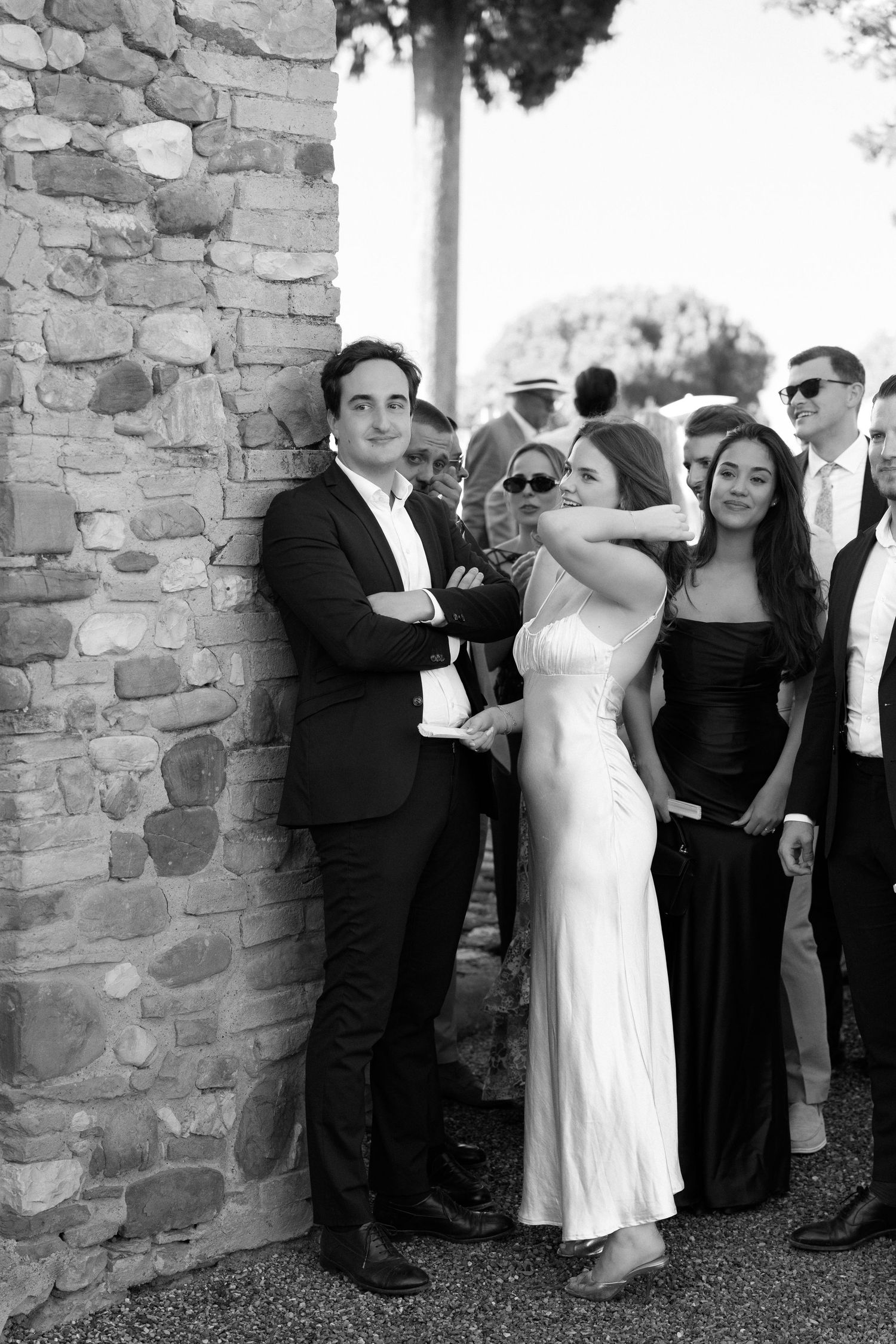 Black and white photo of elegantly dressed wedding guests posing by a stone wall at an outdoor event.
