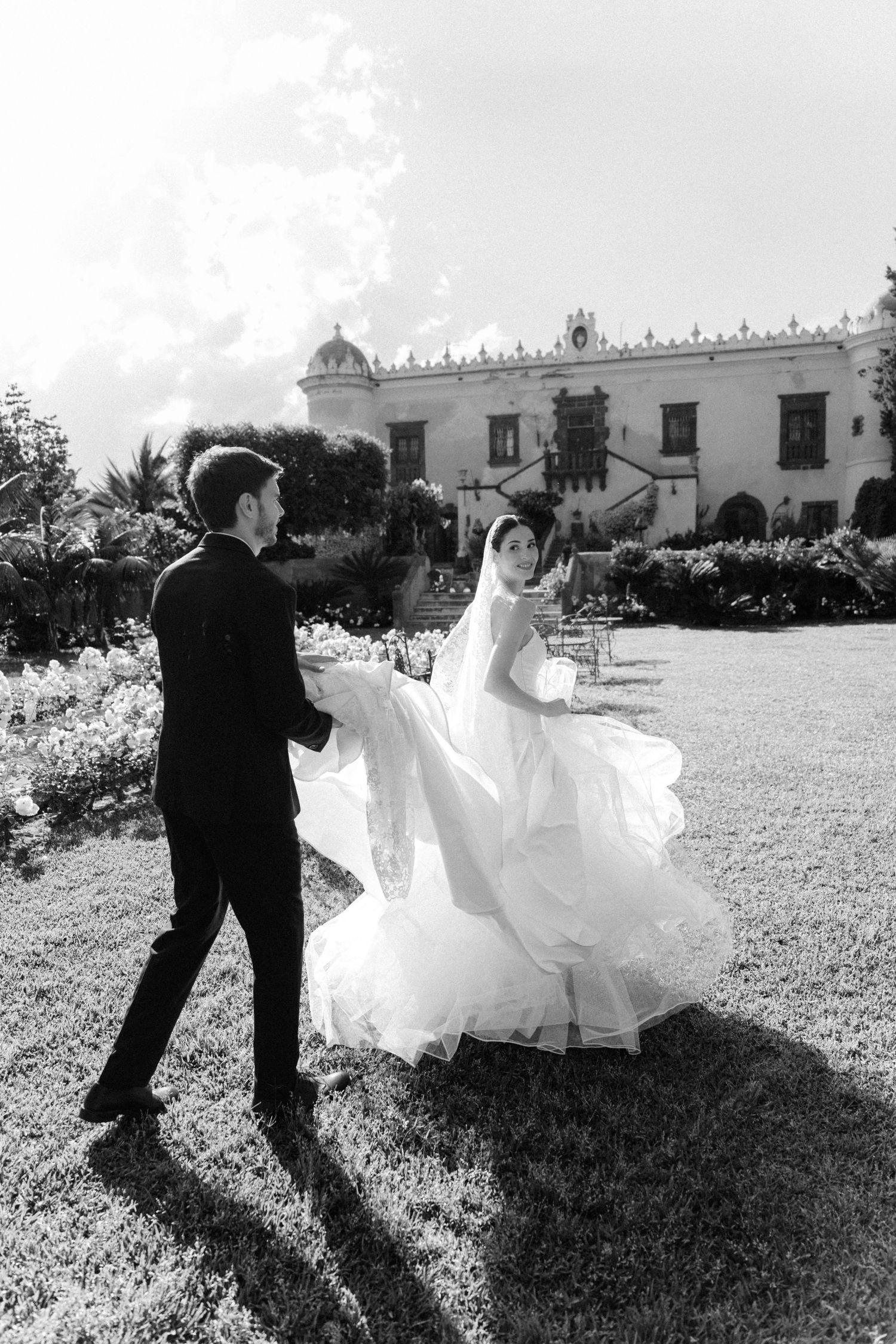 Black and white photo of bride and groom dancing on lawn outside elegant estate on their wedding day.