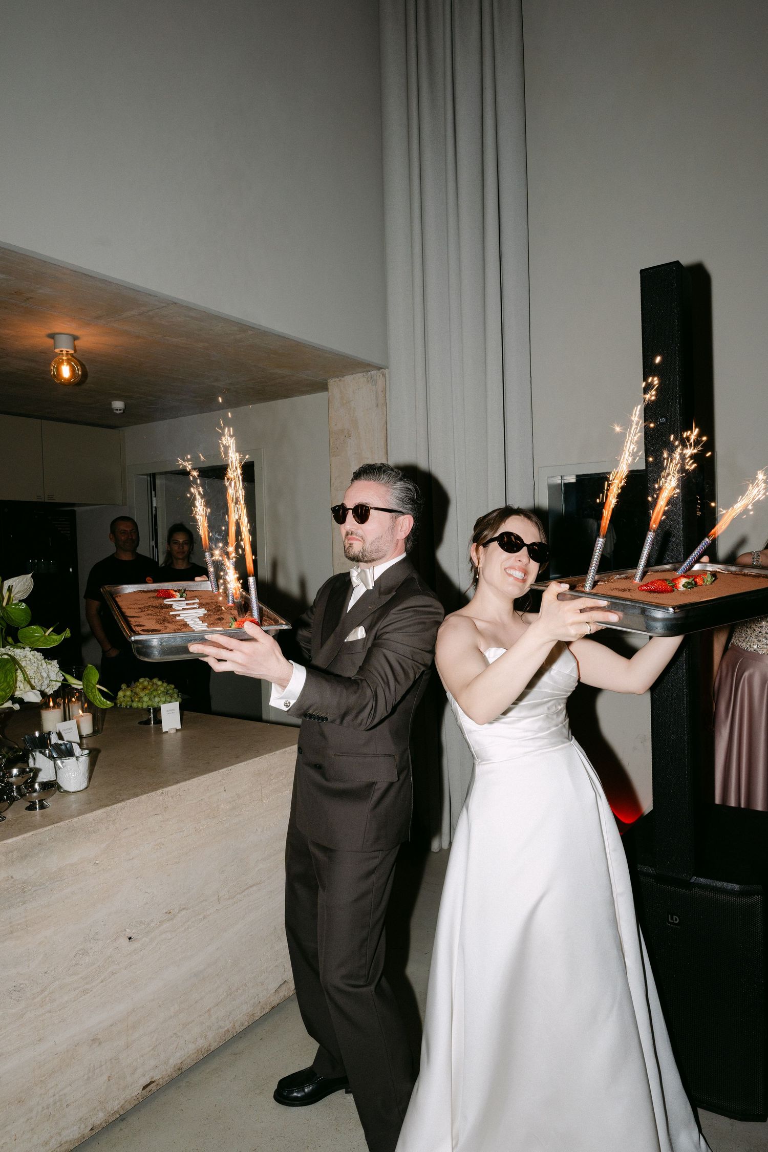 Bride and groom wearing sunglasses hold sparkler trays at their wedding reception indoors.
