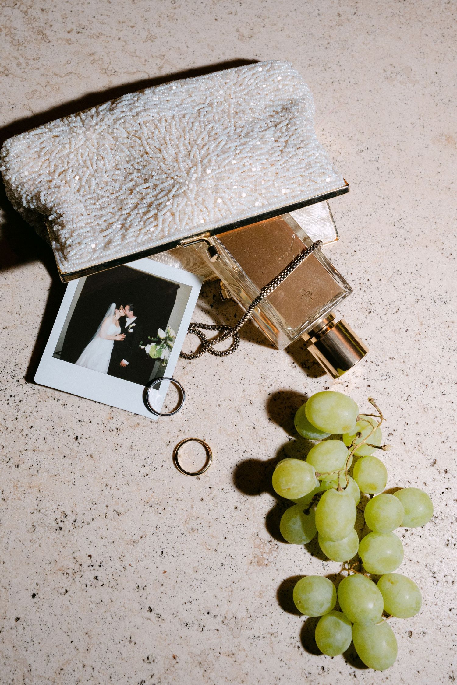 White beaded clutch purse with perfume bottle, polaroid photo, gold ring, and green grapes on beige surface.