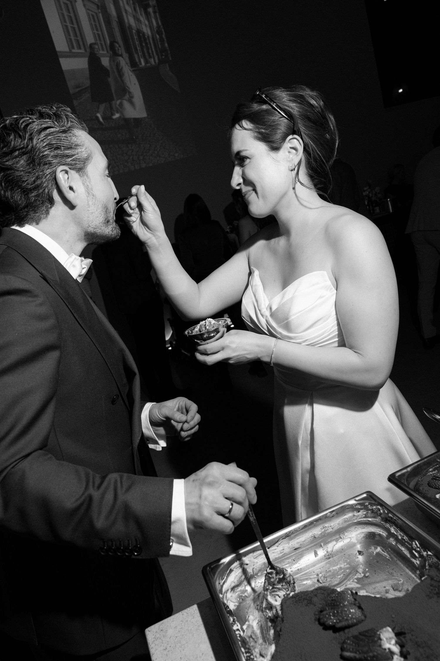 Bride in white gown feeding groom in suit at wedding reception buffet, black and white photo.