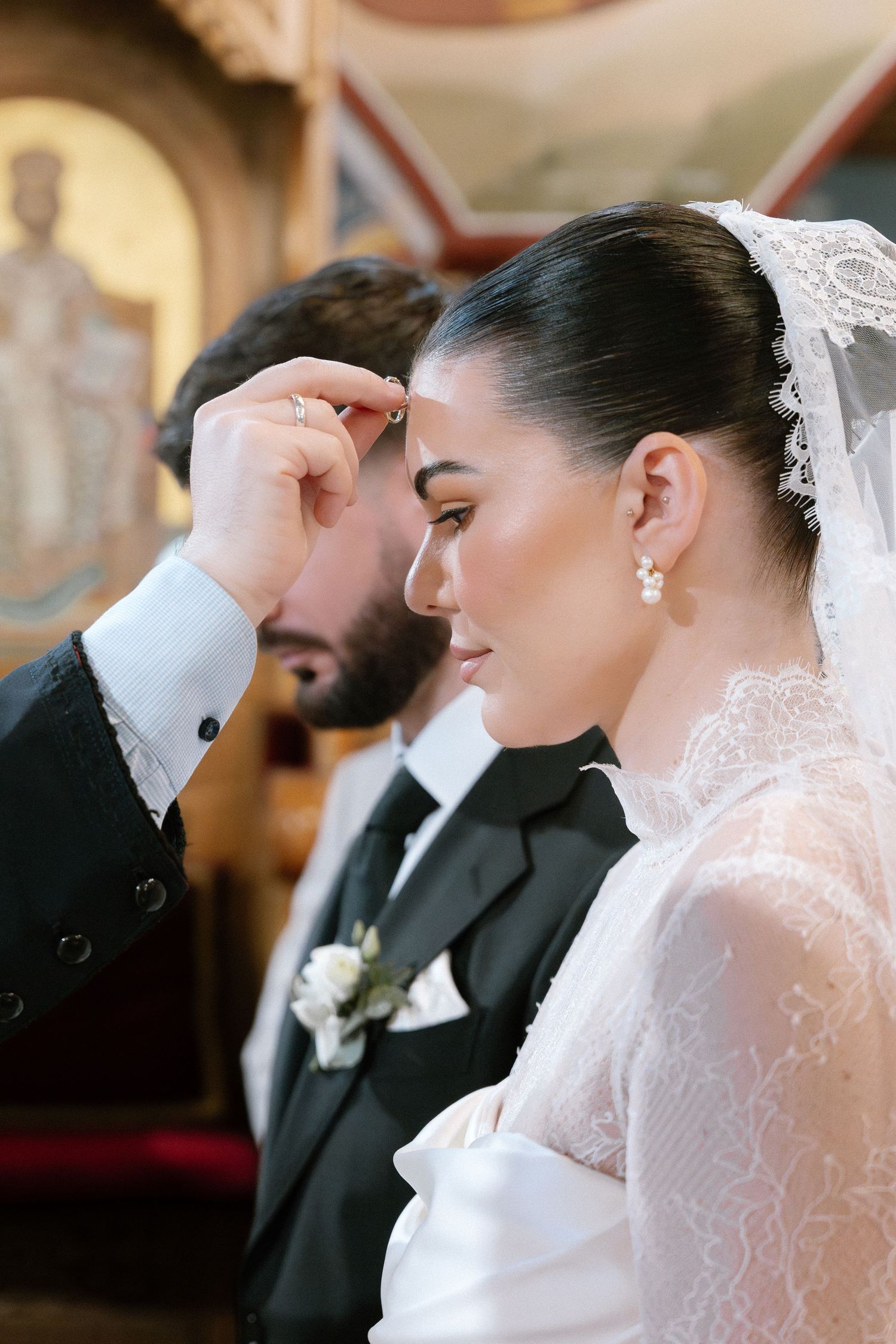 Groom placing wedding ring on brides finger during Orthodox church ceremony, bride wearing lace veil and pearl earrings.