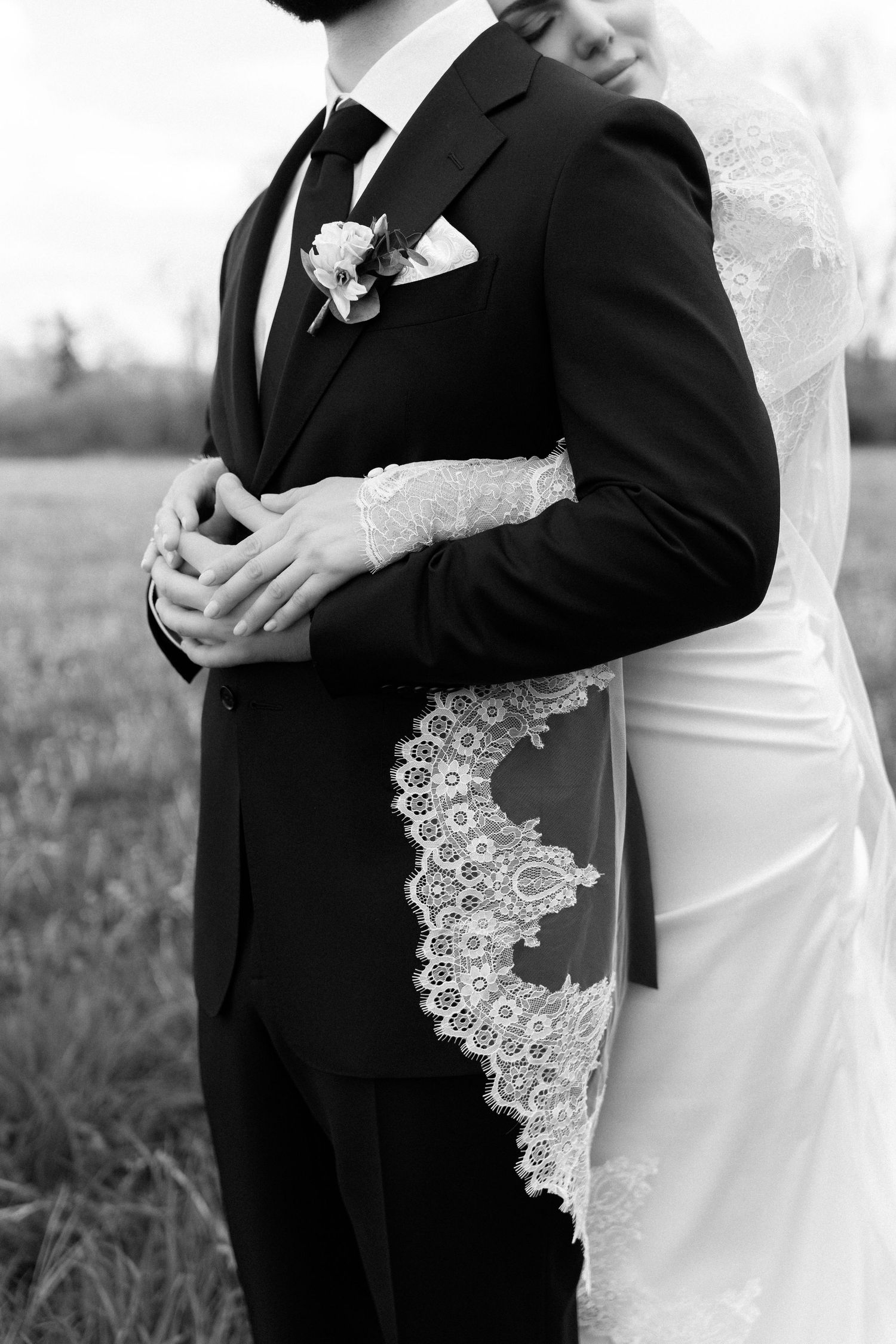 Black and white photo of bride in lace gown embracing groom in black suit with boutonniere at outdoor wedding.