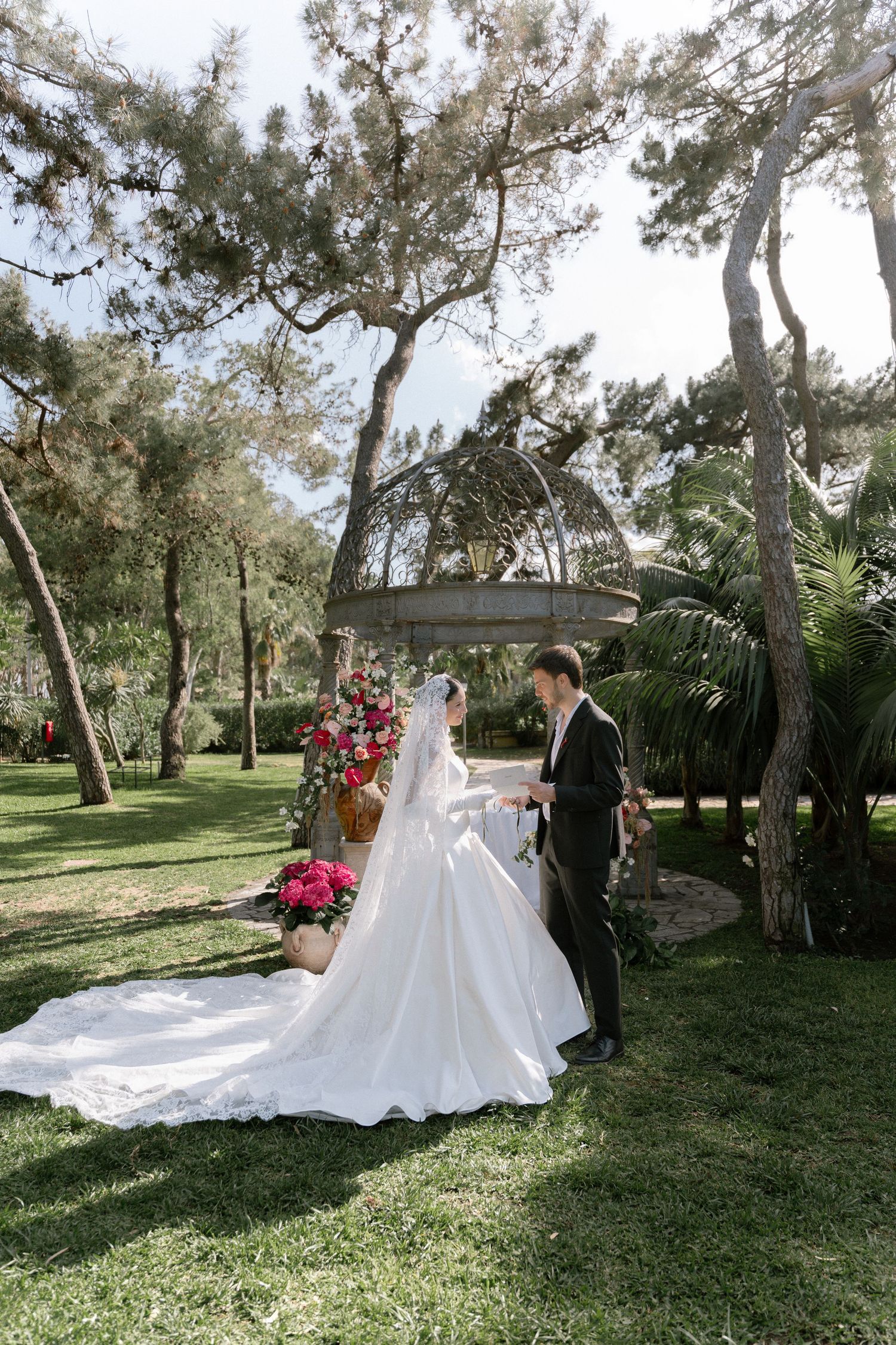 Bride in white gown with long veil and groom in dark suit stand in lush garden near gazebo and pink floral arrangements.