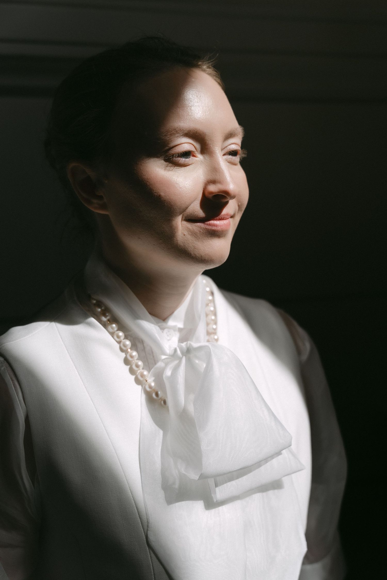 Woman in white blouse with pearl necklace and bow tie smiling softly against dark background.