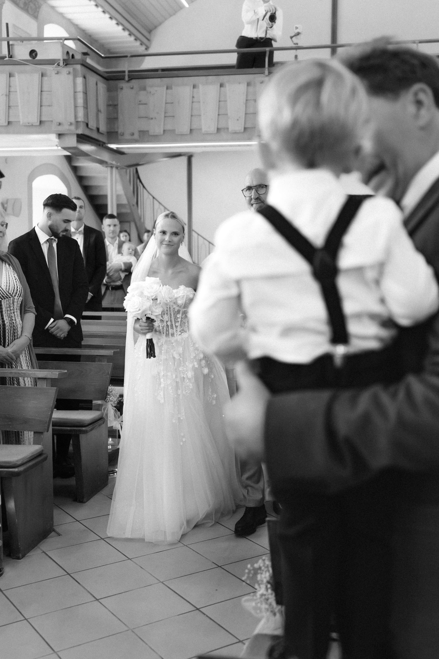 Black and white photo of a bride in a lace wedding gown holding a bouquet as she walks down the church aisle past smiling guests.