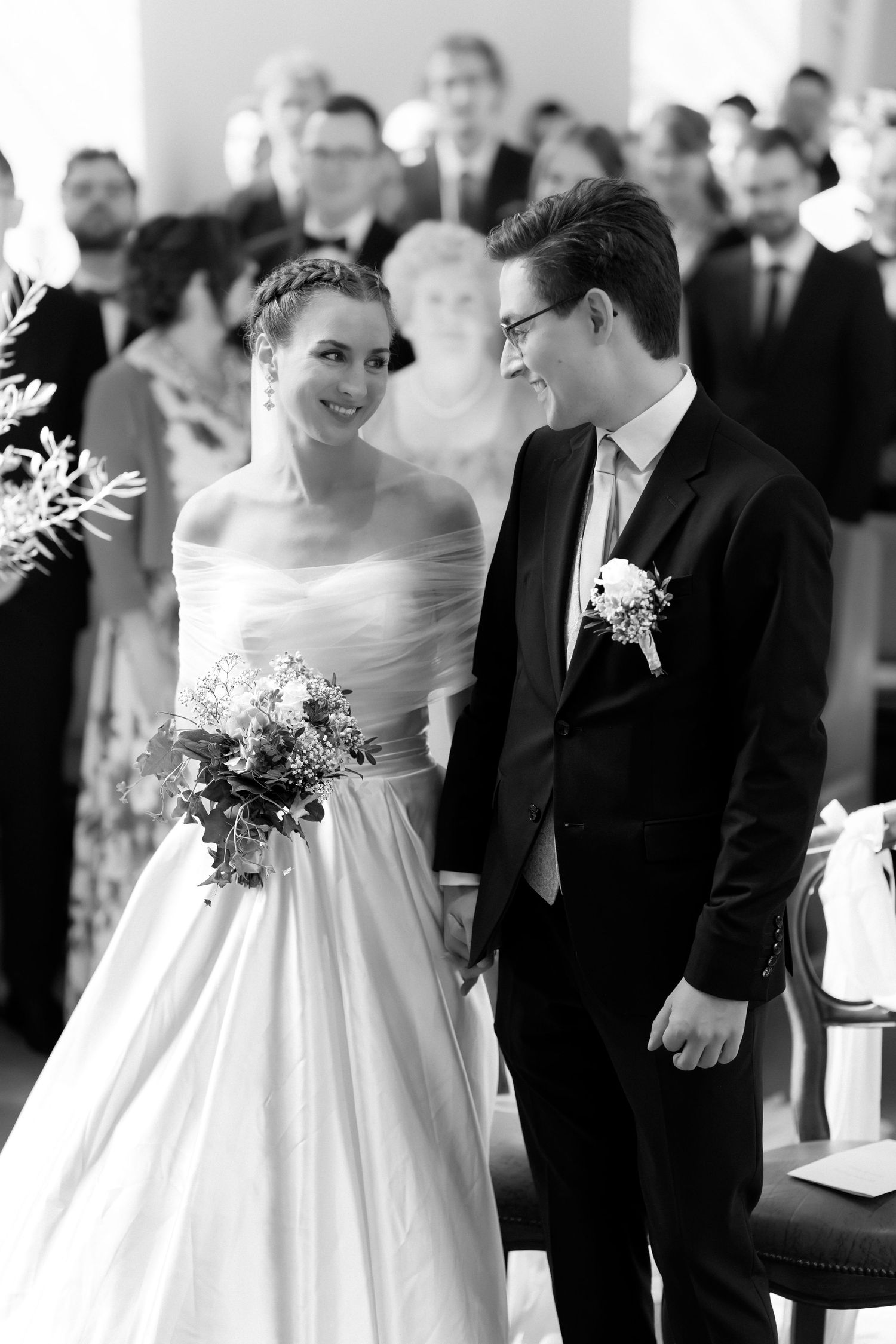 Smiling bride in an off-shoulder satin gown holding a bouquet stands beside a groom in a dark suit during a wedding ceremony with guests.