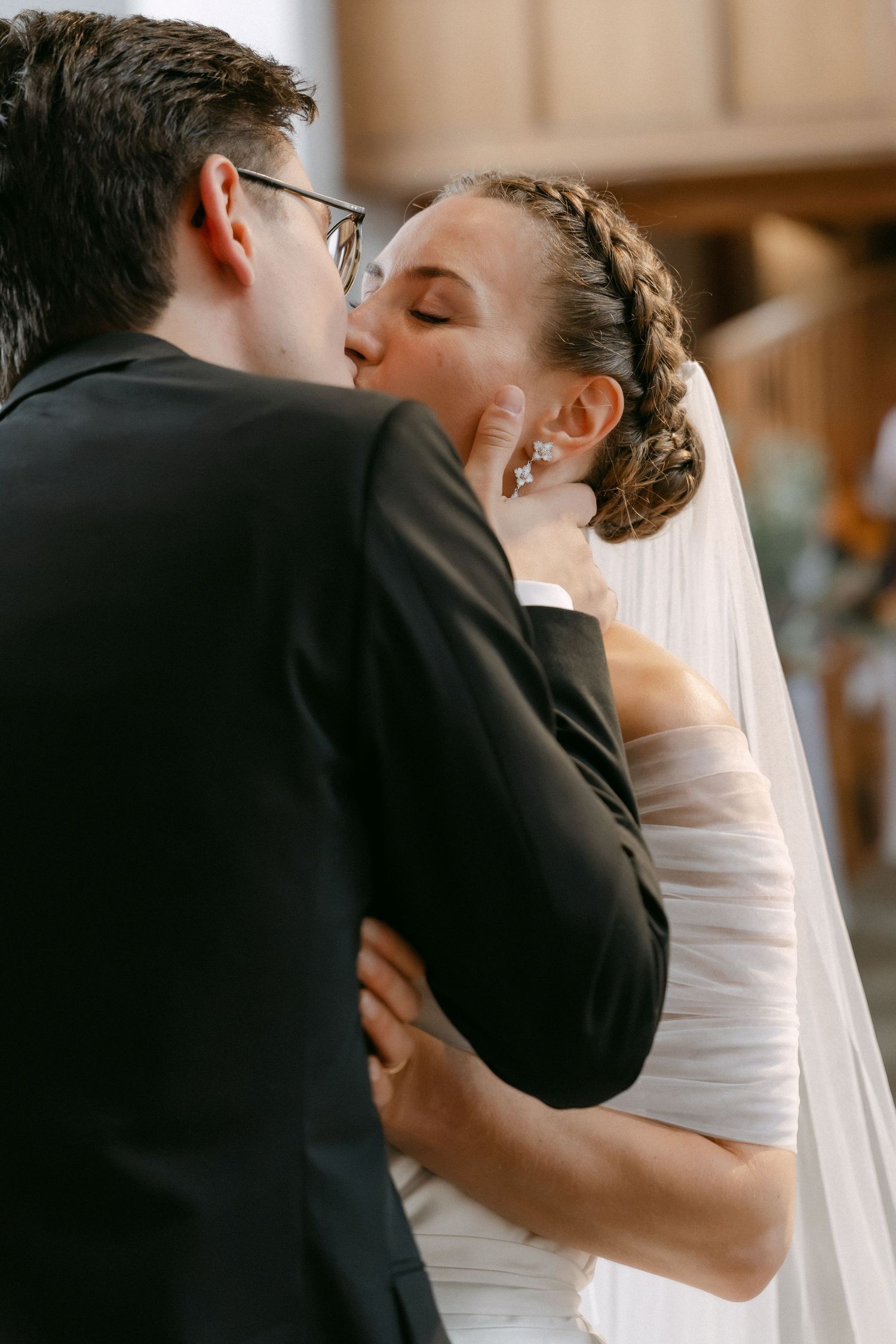 Groom in a dark suit and glasses kisses his bride, who wears a braided updo and a long veil, during a wedding ceremony.