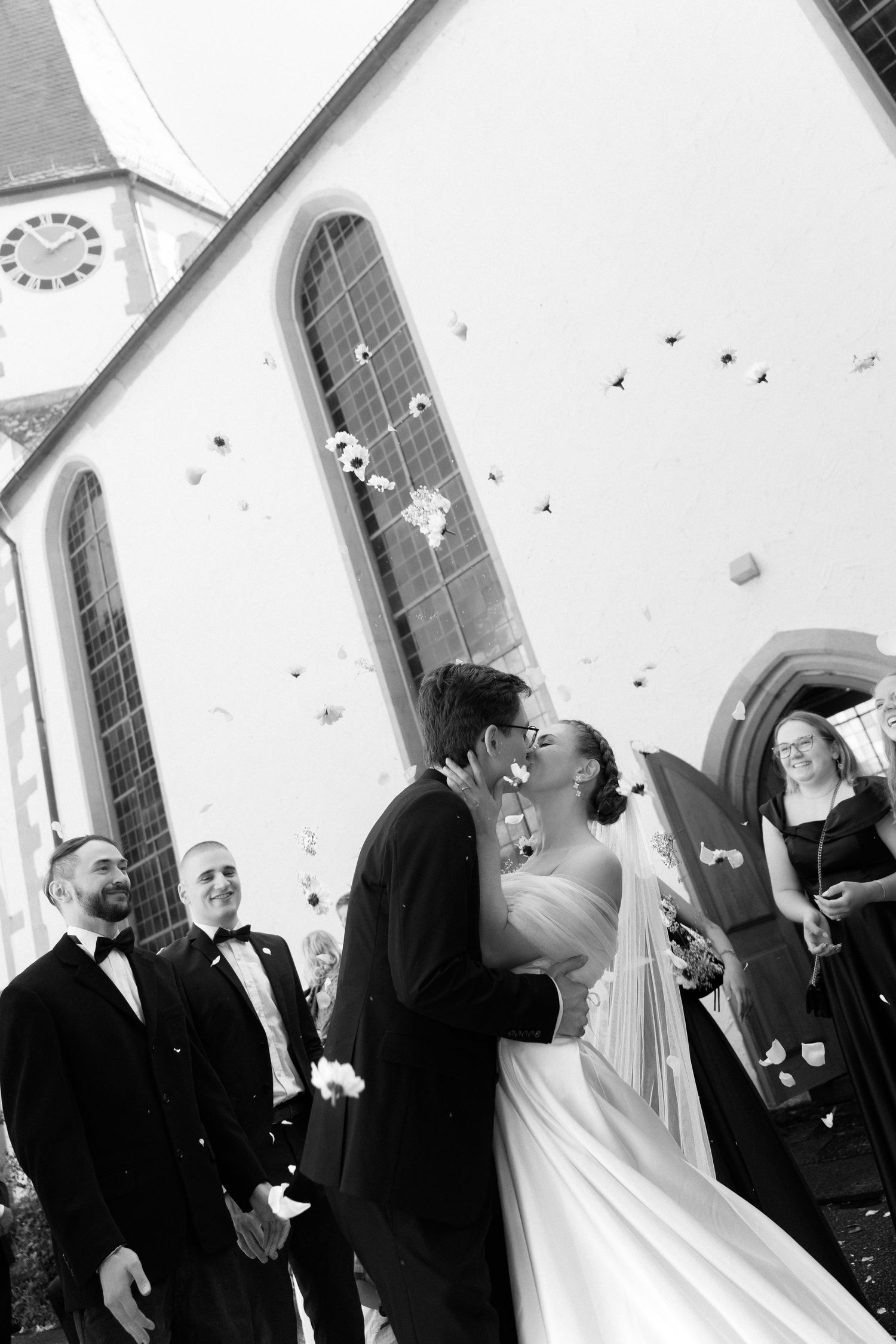 Bride and groom share a kiss outside a church as guests in formal attire watch and confetti falls in this black and white wedding photo.