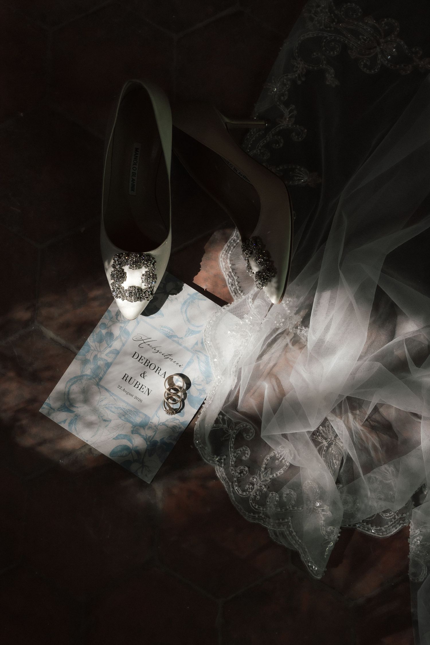 Bridal flat-lay with a jeweled white heel, lace veil, wedding rings and a blue floral invitation card in dramatic light.
