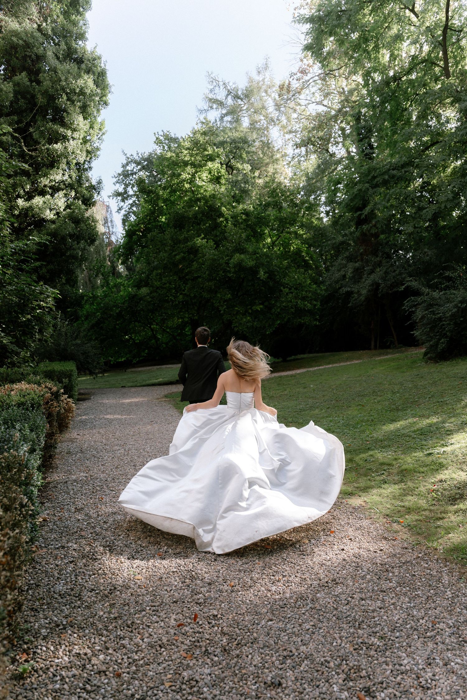 Bride in a flowing white strapless gown runs down a gravel path toward a groom in a dark suit, surrounded by lush green trees.