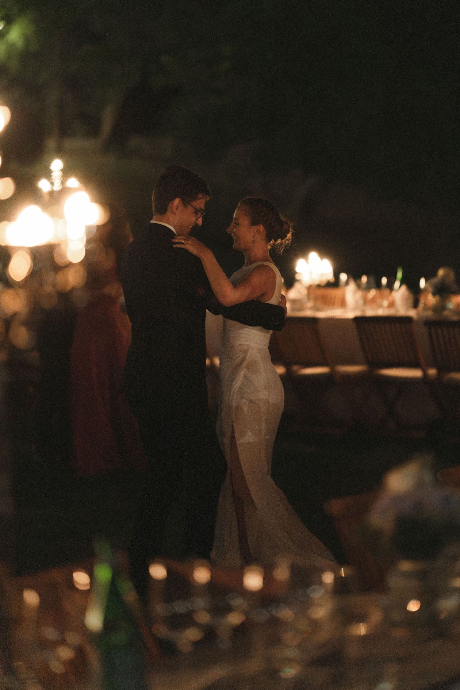 A couple in formal wear shares a slow dance at an outdoor evening wedding reception, with glowing candlelit tables in the background.