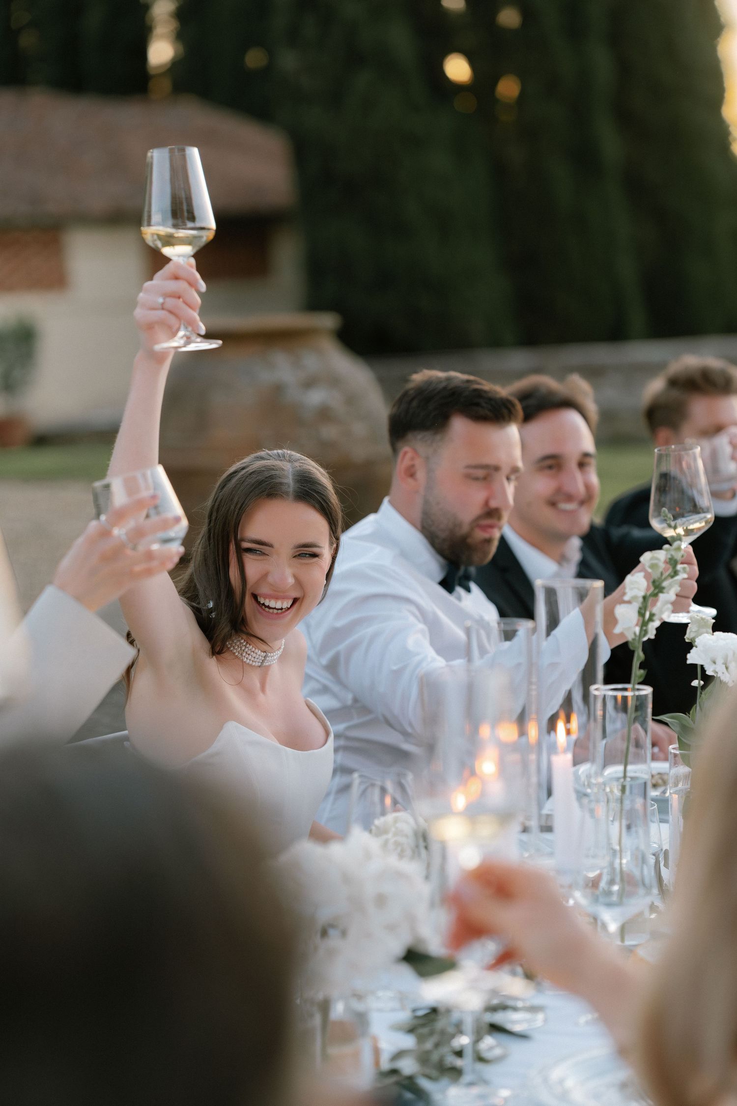 Bride raises champagne glass joyfully at outdoor wedding reception dinner table with guests celebrating.