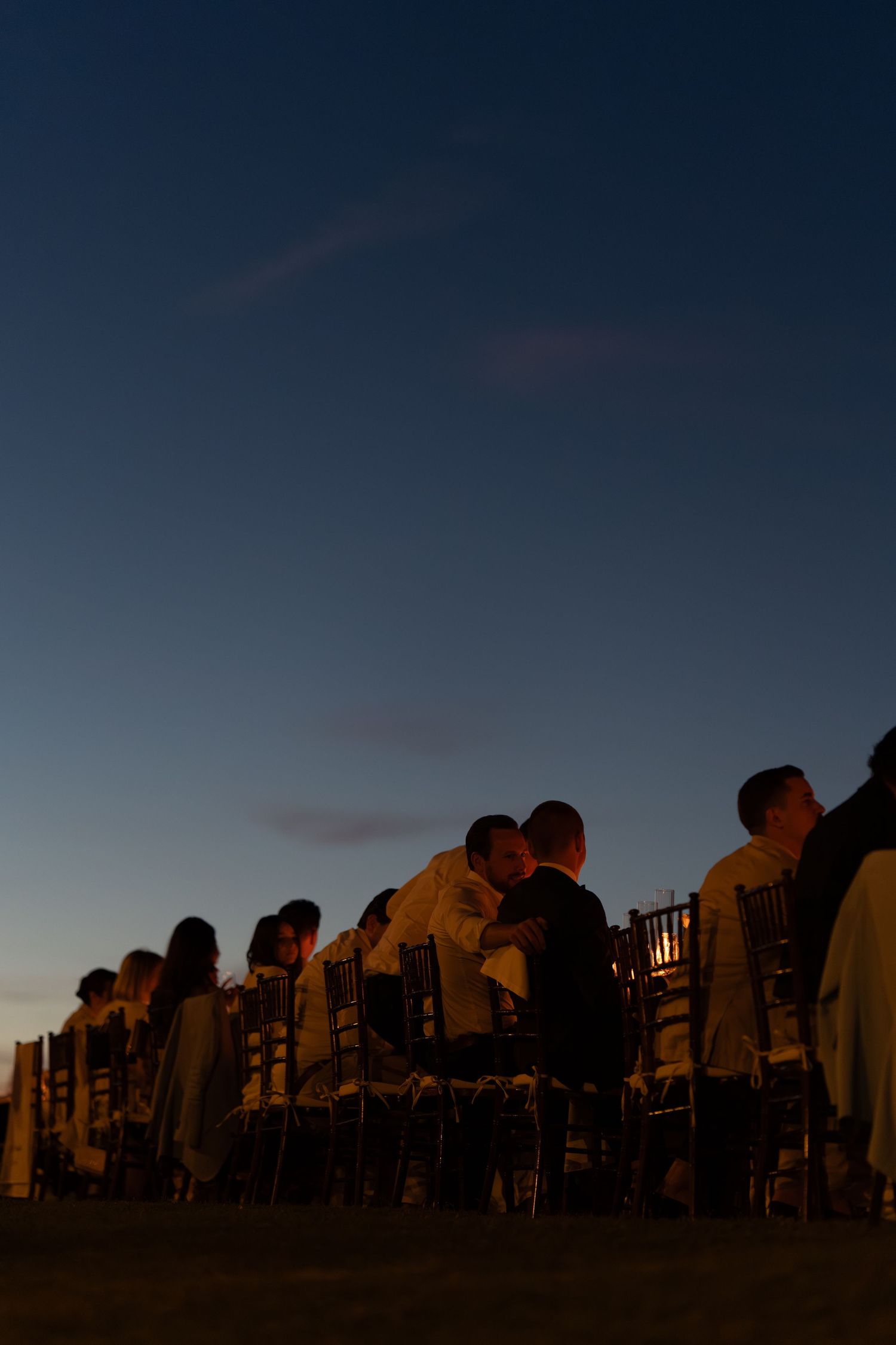 Silhouetted wedding guests seated in chairs outdoors at dusk, with a deep blue twilight sky in the background.