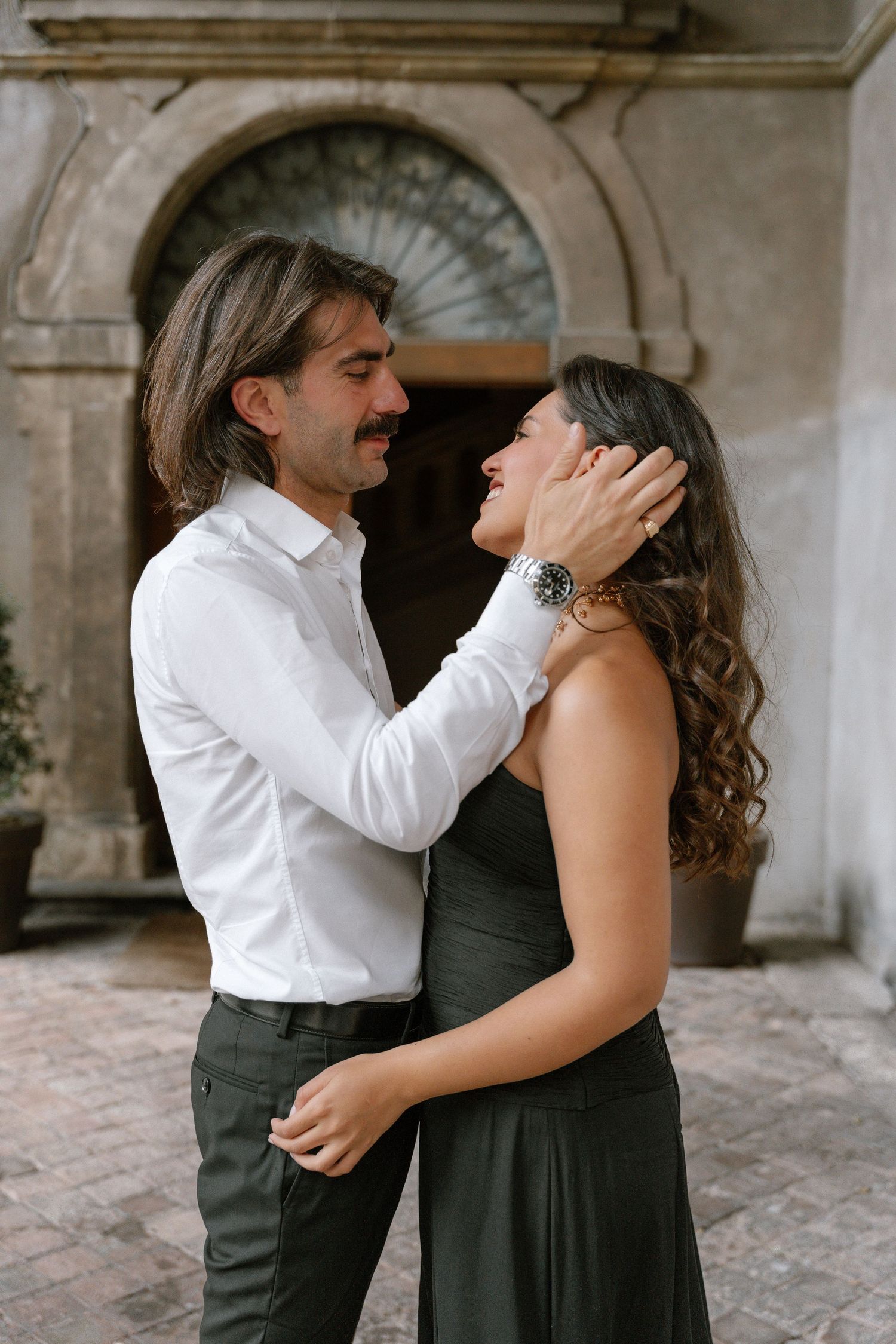 A couple shares a romantic moment, man in white shirt gently holds woman's face, both smiling, against stone building.