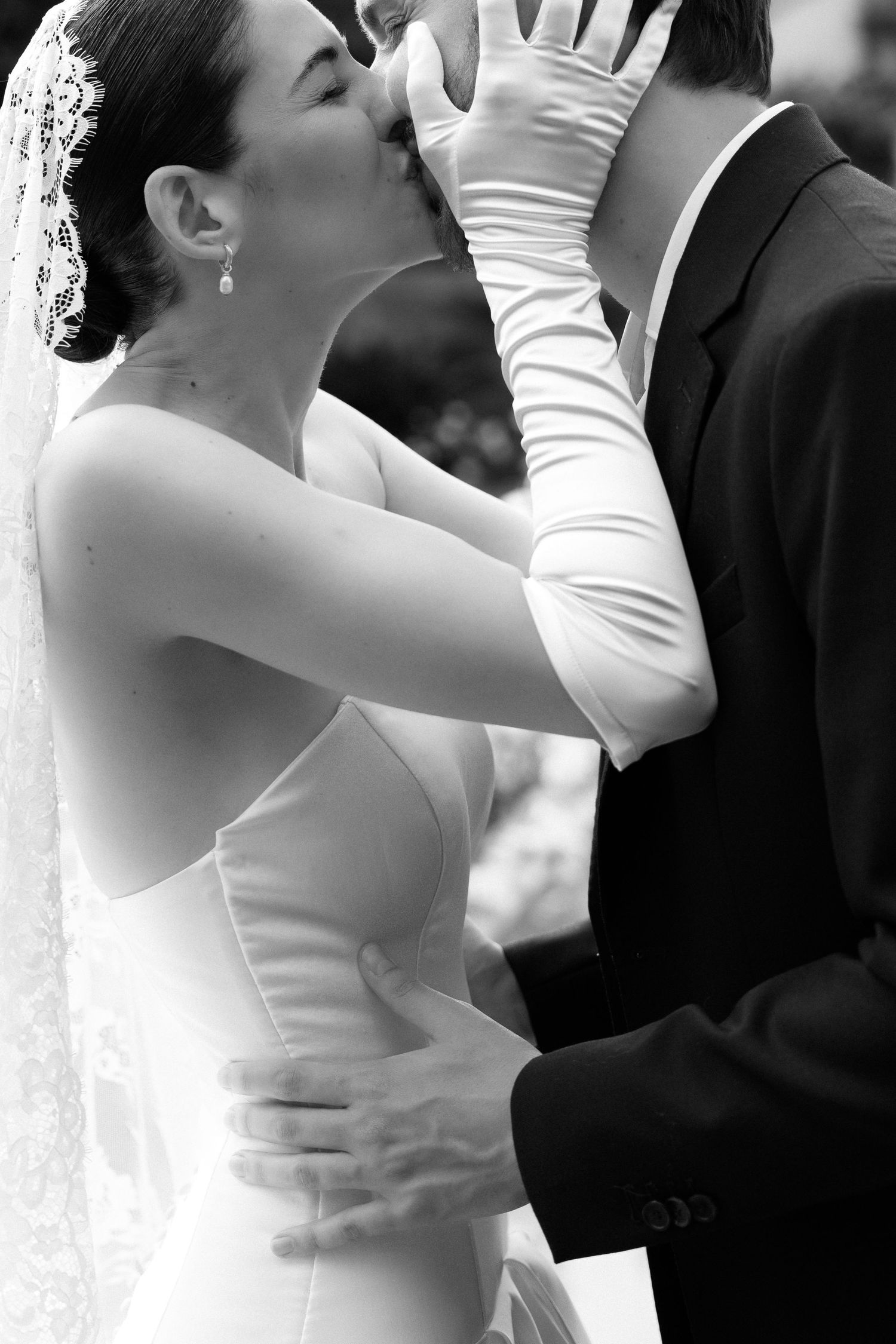 Black and white photo of a bride in white gown and gloves sharing a kiss with groom in suit at wedding.