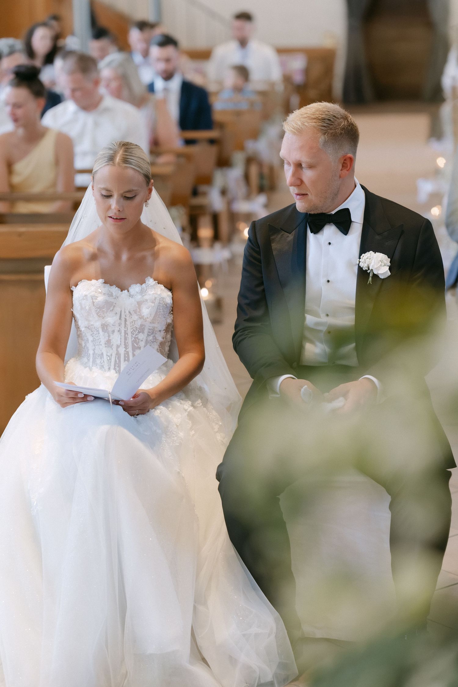 Bride in a strapless lace wedding gown and veil reads her vows beside a groom in a black tuxedo and bow tie during a church ceremony.