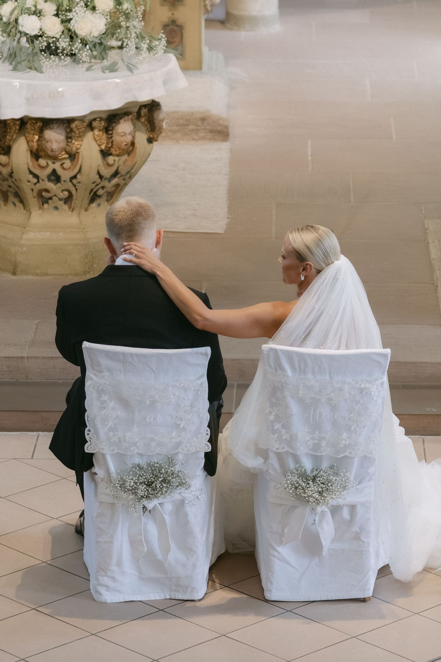 Bride in a white veil rests her hand on the groom's shoulder as the couple sit on decorated chairs during a church wedding ceremony.