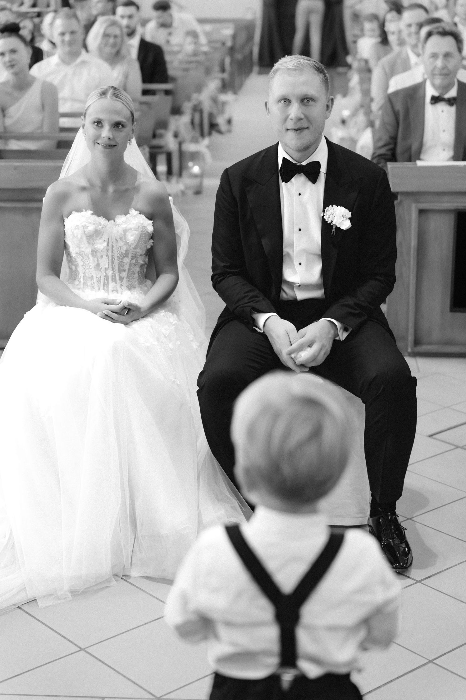Black and white photo of a bride and groom seated during a church wedding ceremony as a young ring bearer in suspenders faces them.