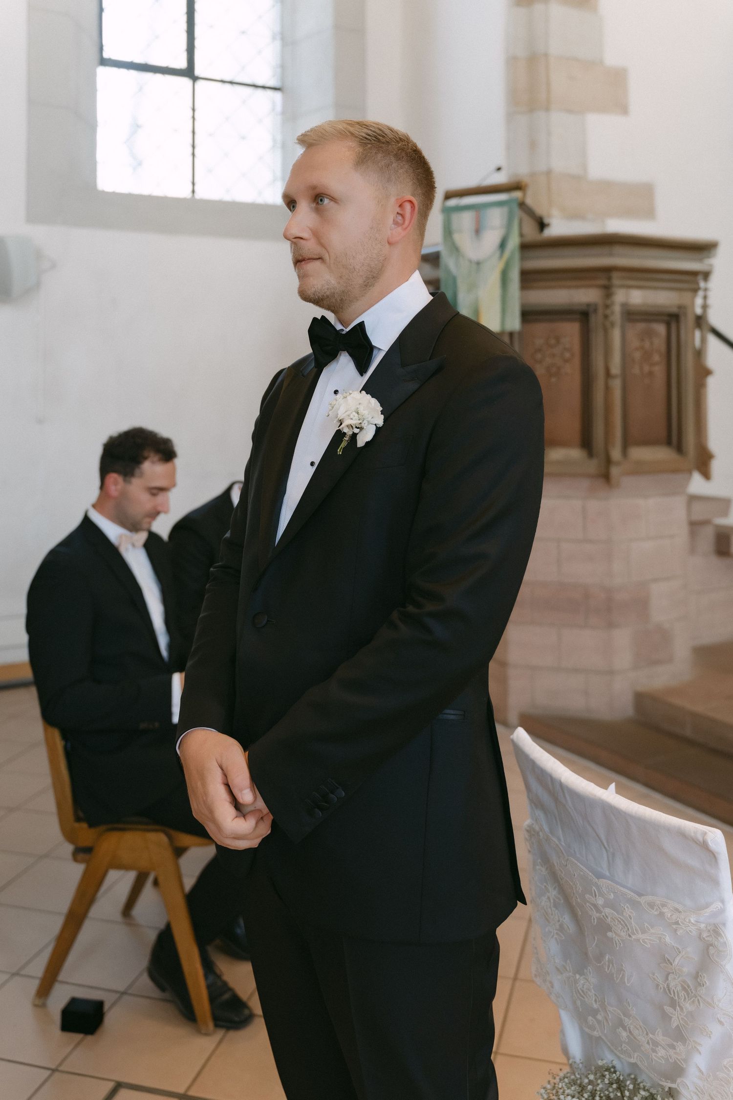Groom in a black tuxedo with bow tie and white boutonniere stands at the altar of a stone church while a seated man looks on.