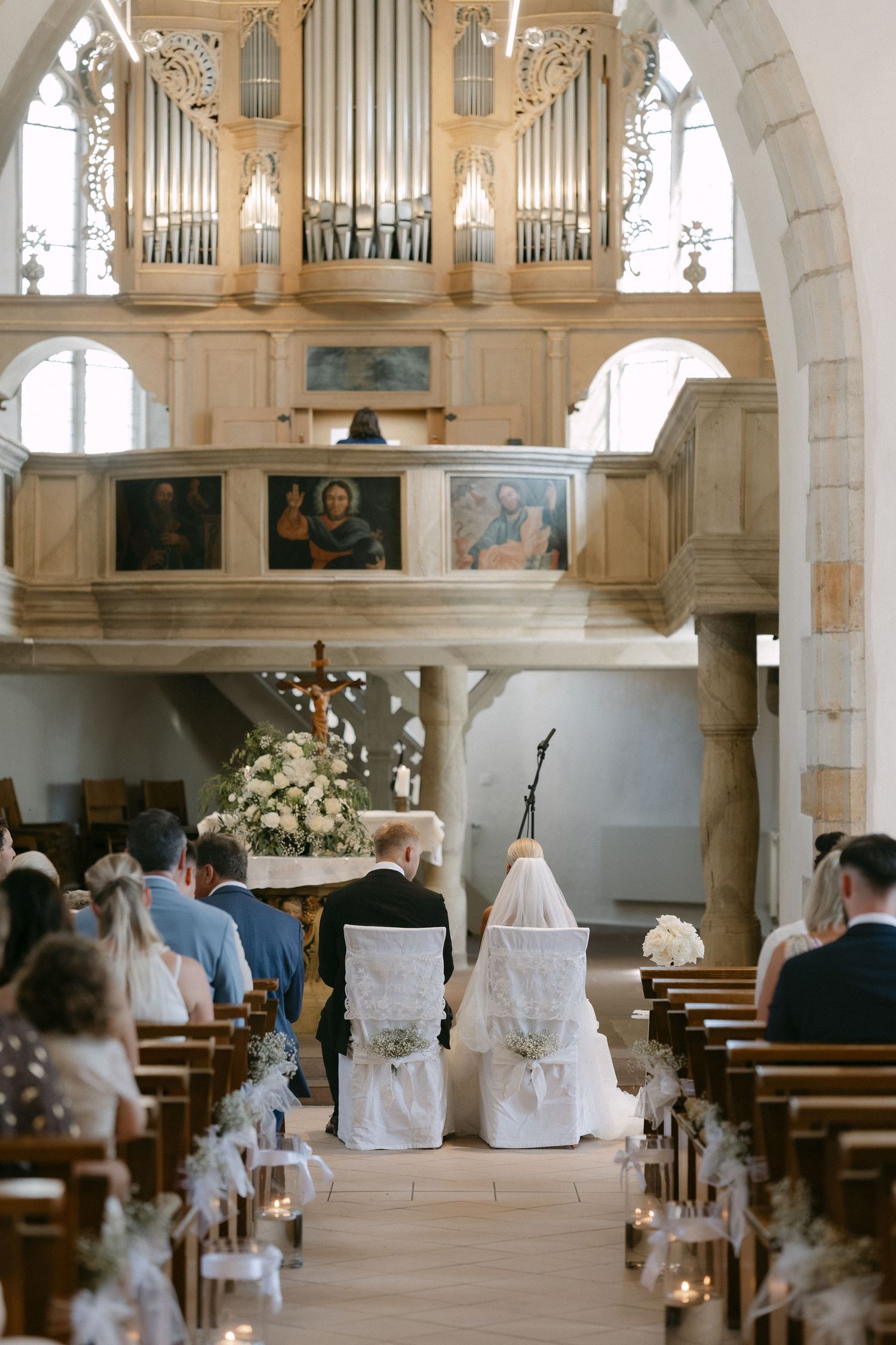 Bride and groom stand before the altar during a church wedding ceremony, with guests seated in pews and an ornate pipe organ above.
