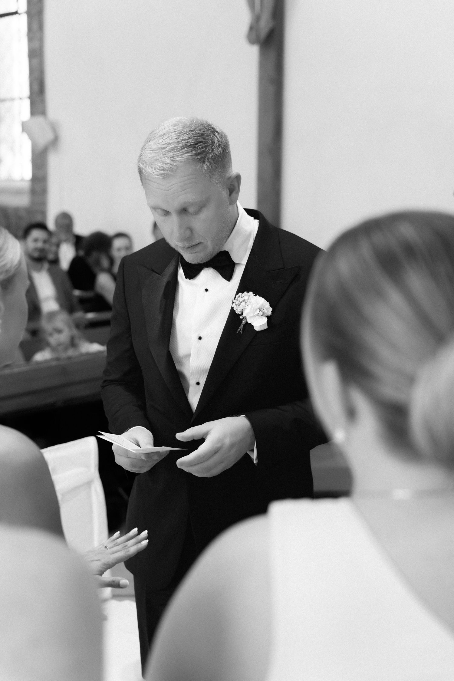 Groom in a black tuxedo with bow tie and floral boutonniere reads from a card during a wedding ceremony as guests look on.