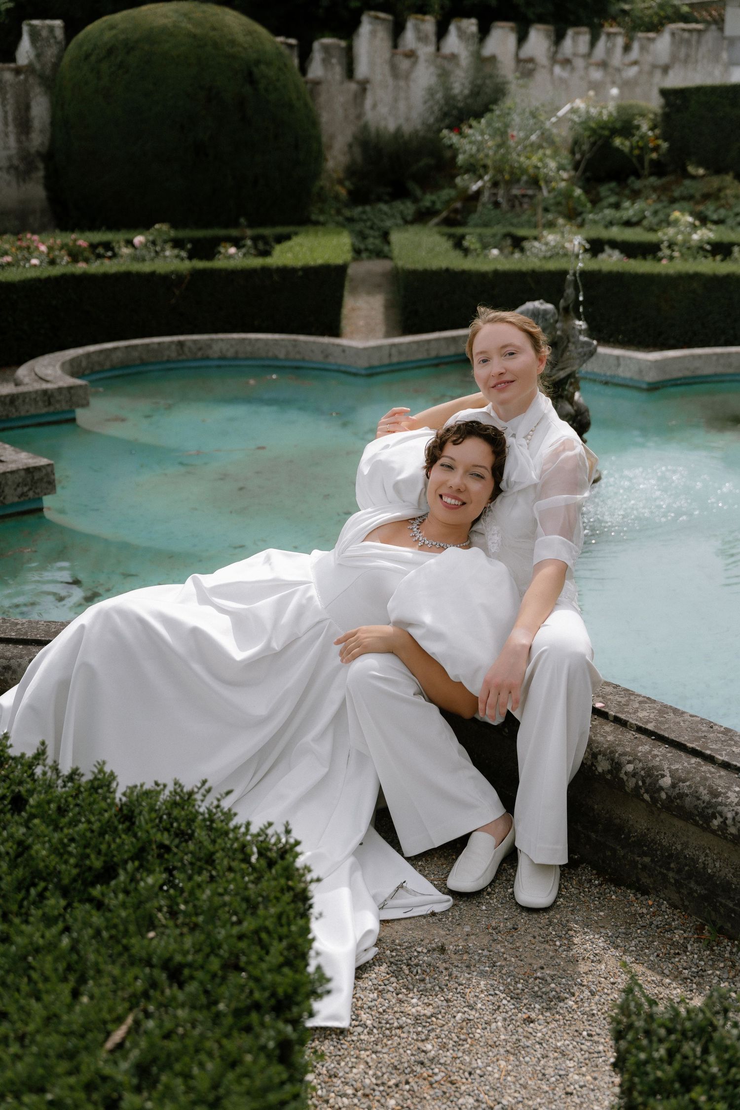 Two brides in white wedding attire pose elegantly by a garden fountain surrounded by lush greenery.