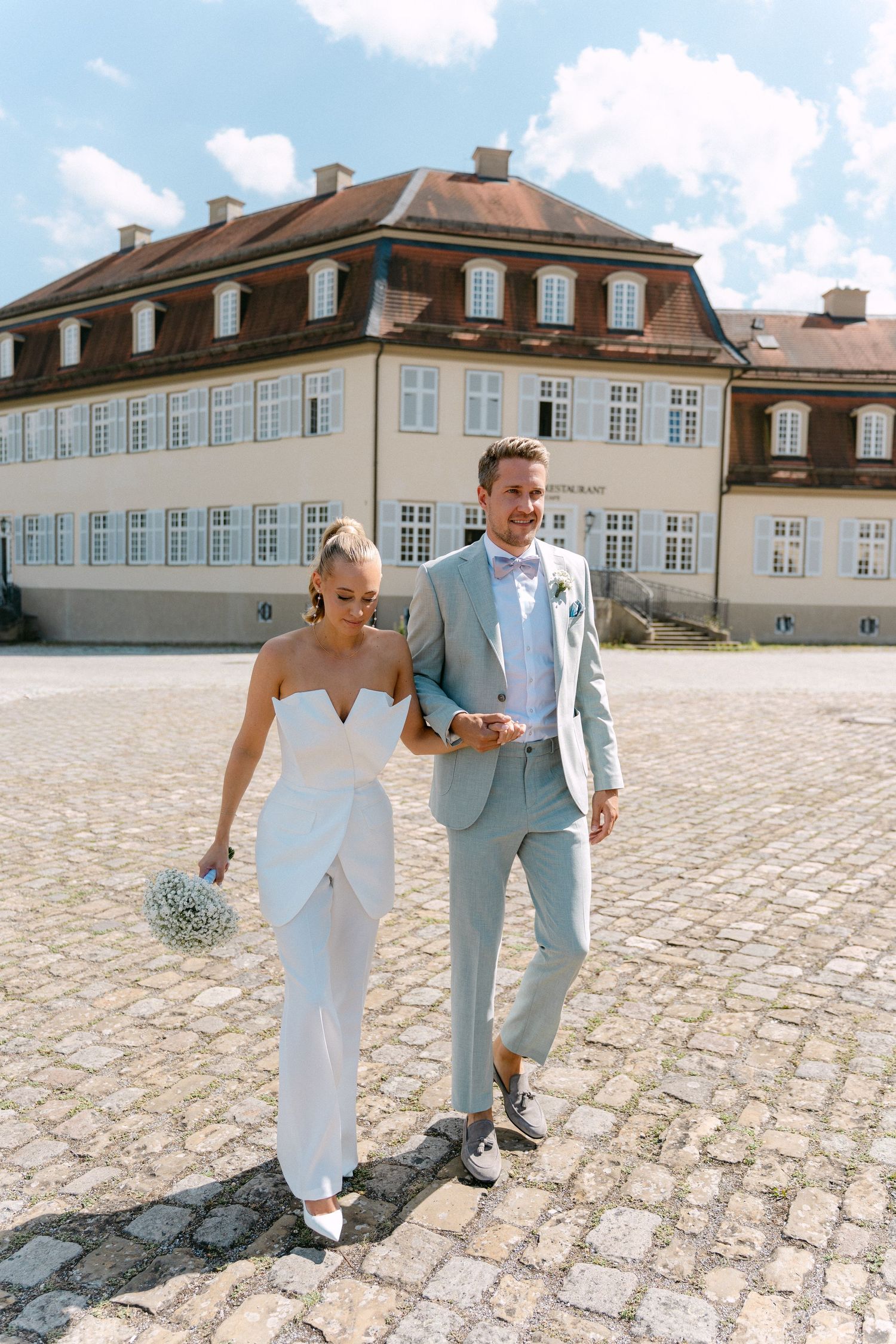 A couple in wedding attire walks and laughs together on a cobblestone courtyard outside a historic mansion. Schloss Solitude